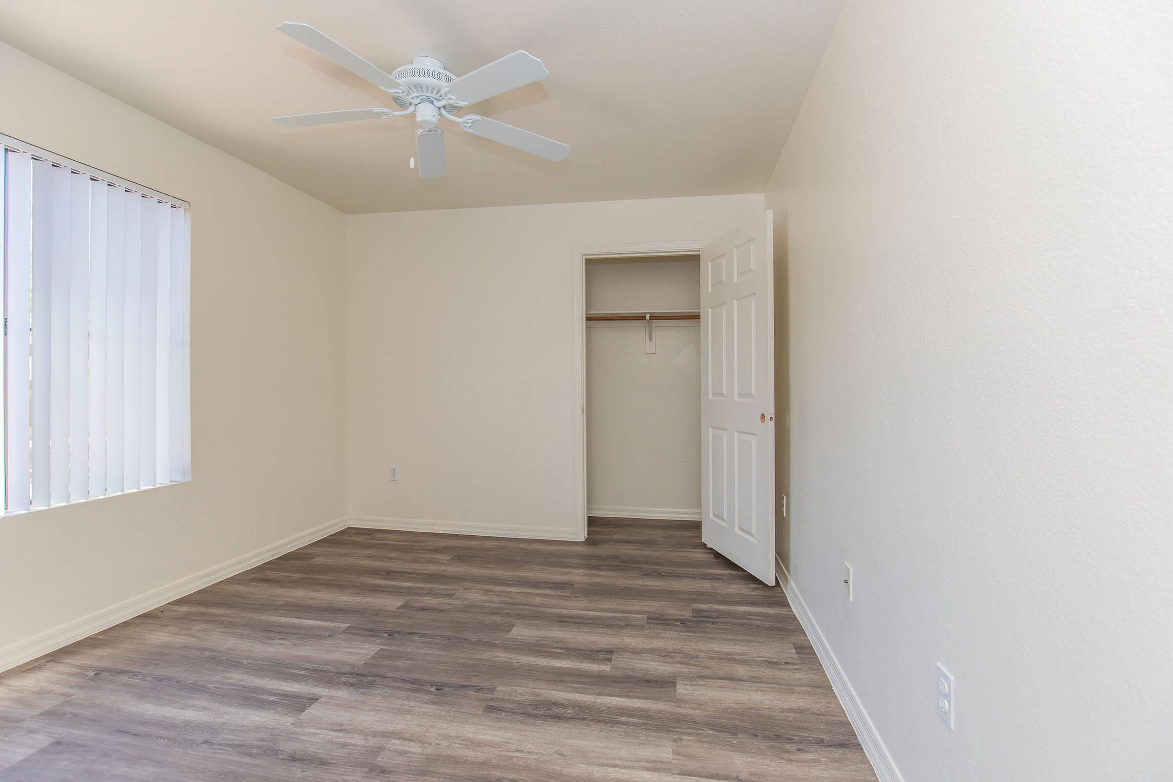 A simple, empty room with light-colored walls and a ceiling fan. The floor features light wood laminate, and there is a closet with a door slightly open. A window with blinds provides natural light, creating a spacious and airy atmosphere.