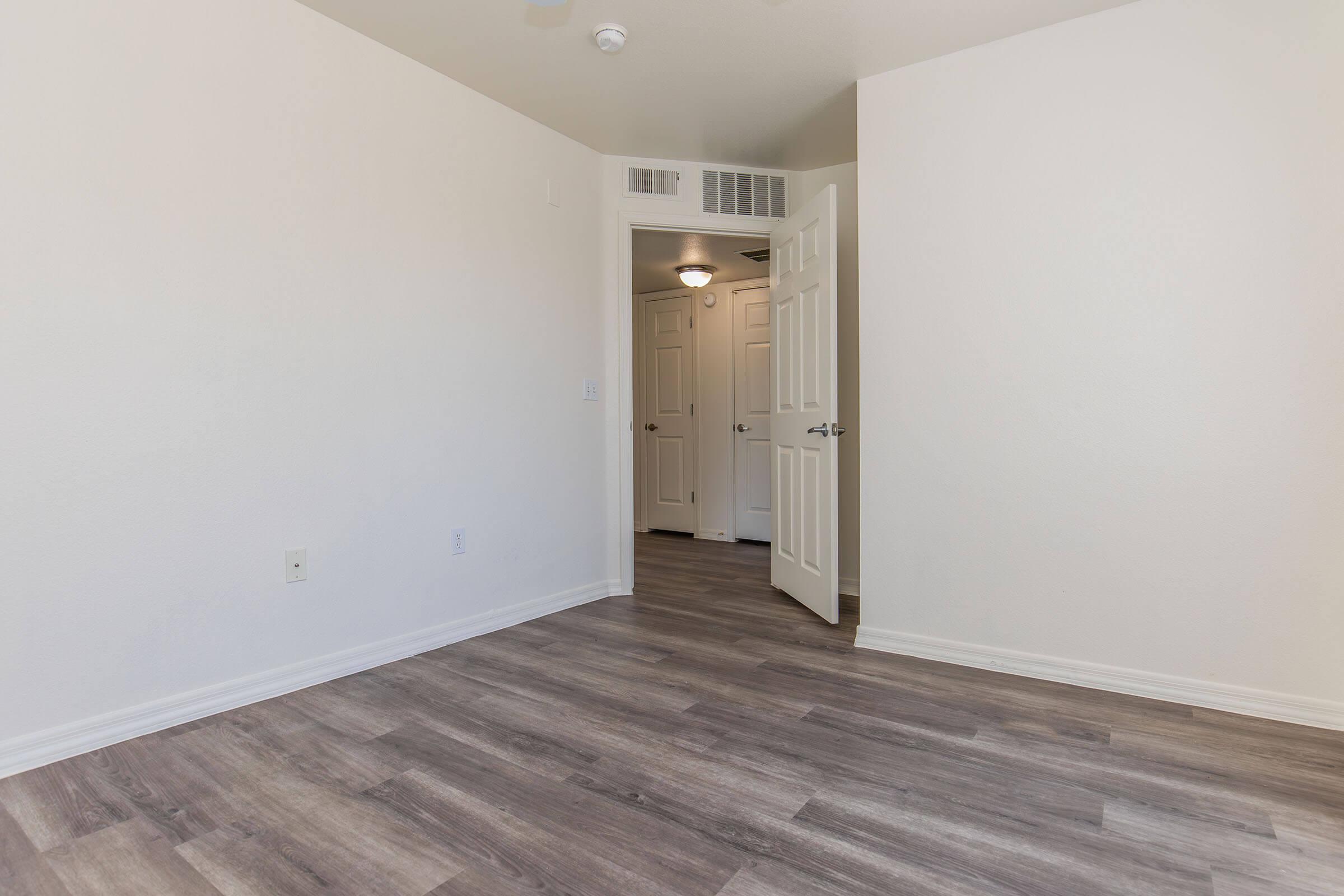 A spacious empty room with light-colored walls and laminate flooring. A partially open door leads to another area, revealing additional doors. The ceiling has a vent, and the room is well-lit by natural light coming from an unseen window.