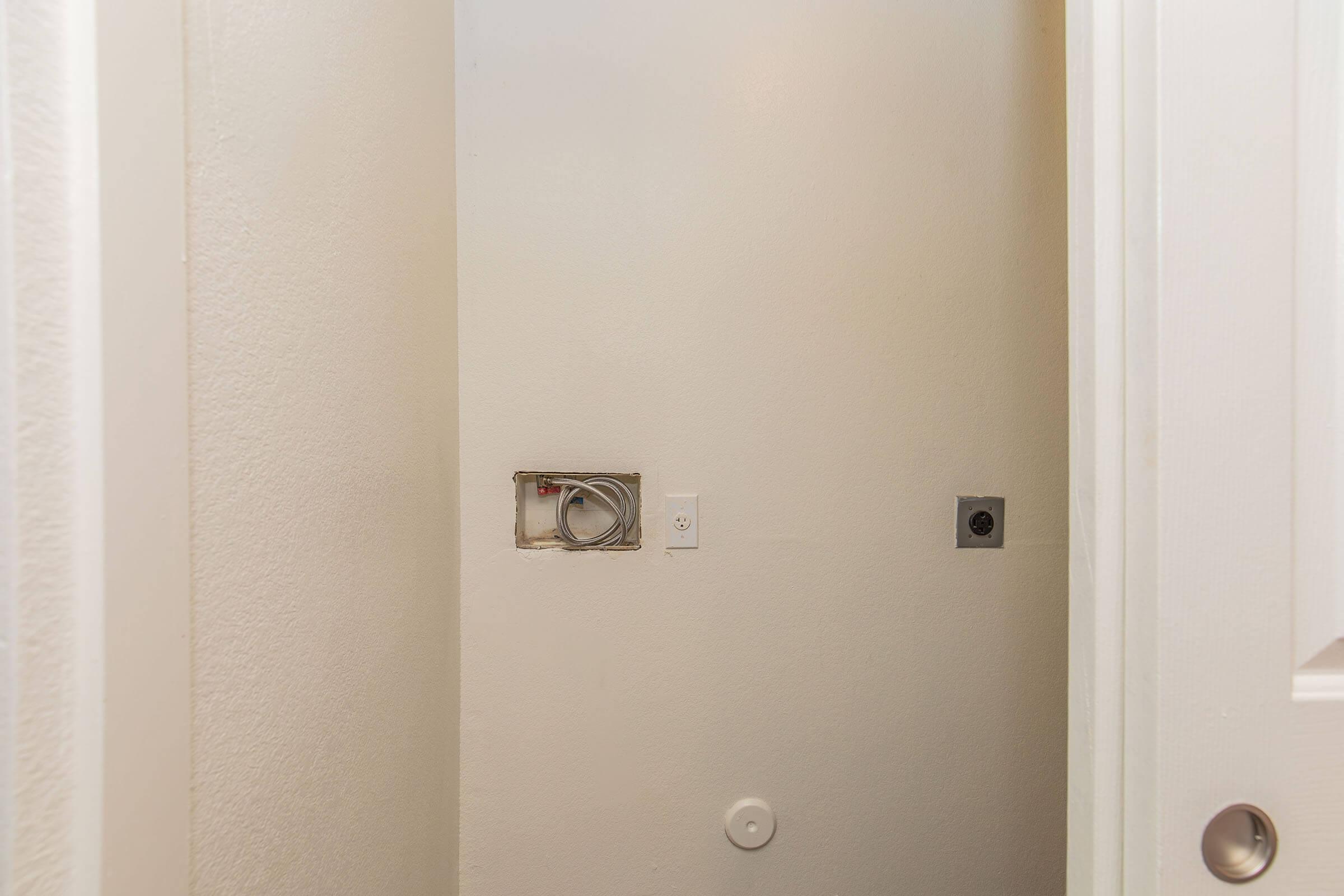 A small, empty room with plain white walls. There are two electrical outlets and a rectangular cut-out in the wall, indicating potential plumbing or wiring. The floor is bare, and the room is accessed through an open door. Overall, the space appears to be unfinished or prep work for an installation.