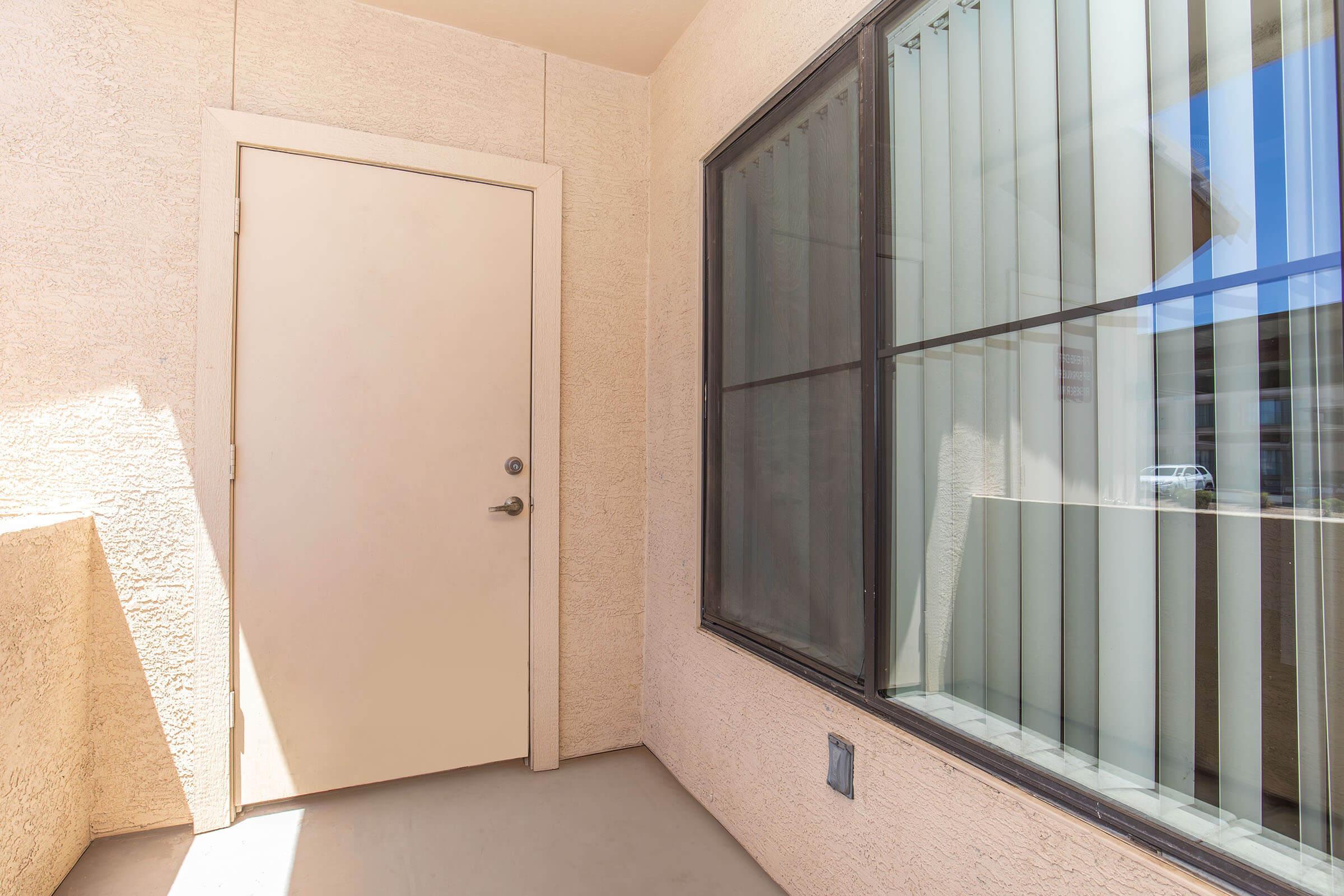 A corner view of an apartment entrance featuring a plain beige door and a large window with vertical blinds. The wall and floor are light-colored, creating a calm atmosphere. Sunlight streams in, highlighting the simplicity of the entrance area.