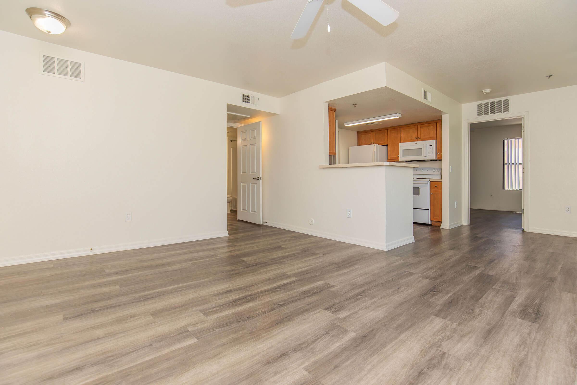A spacious, well-lit apartment interior featuring light-colored walls and laminate flooring. The room includes a ceiling fan and an open floor plan leading to a kitchen area with wooden cabinets and appliances. Doorways lead to additional rooms, creating a modern and inviting living space.