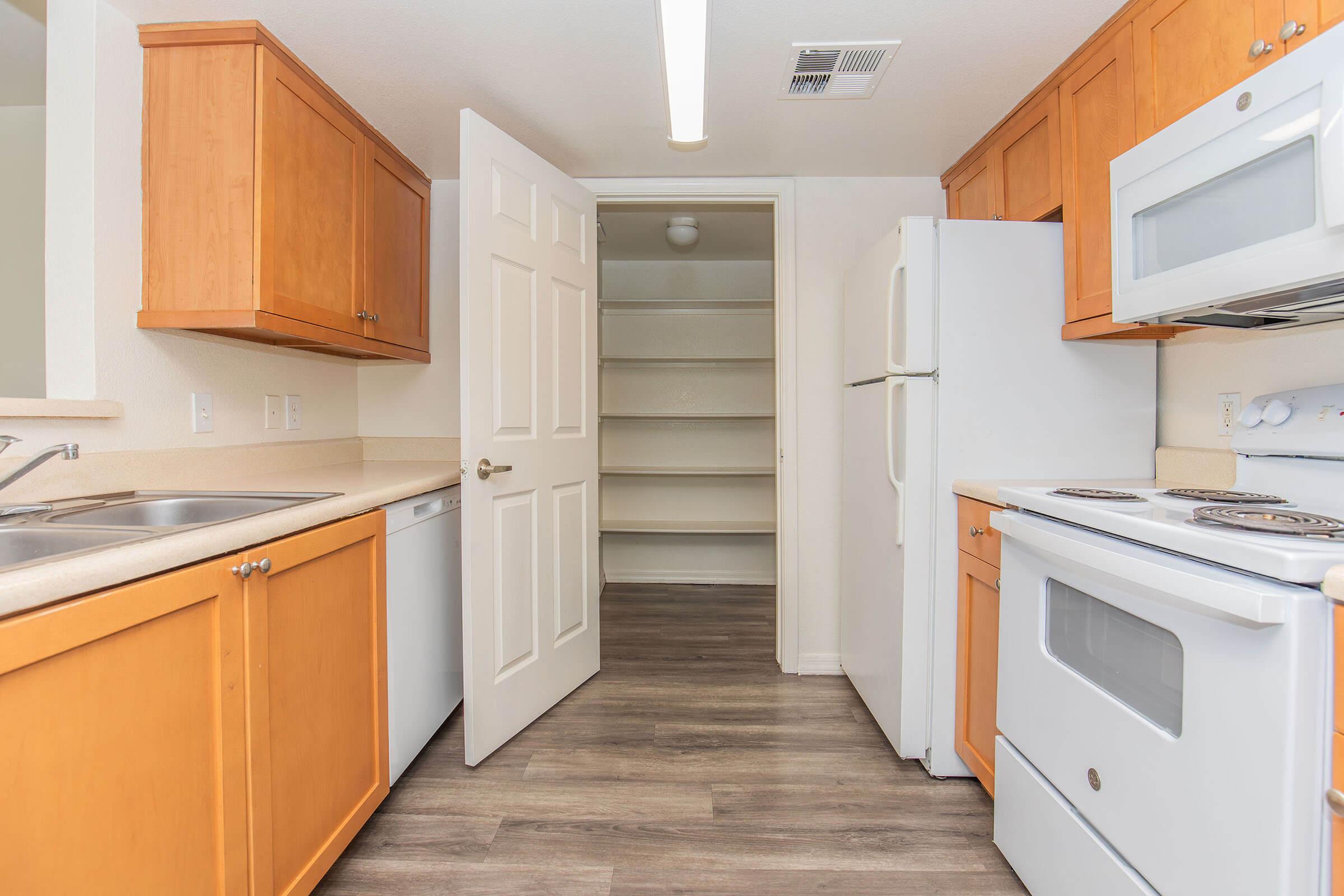 A well-lit kitchen featuring wooden cabinets, a sink, dishwasher, and appliances including a white refrigerator and stove. A door leads to a pantry with shelves on the right. The flooring is a light wood, creating a warm atmosphere.