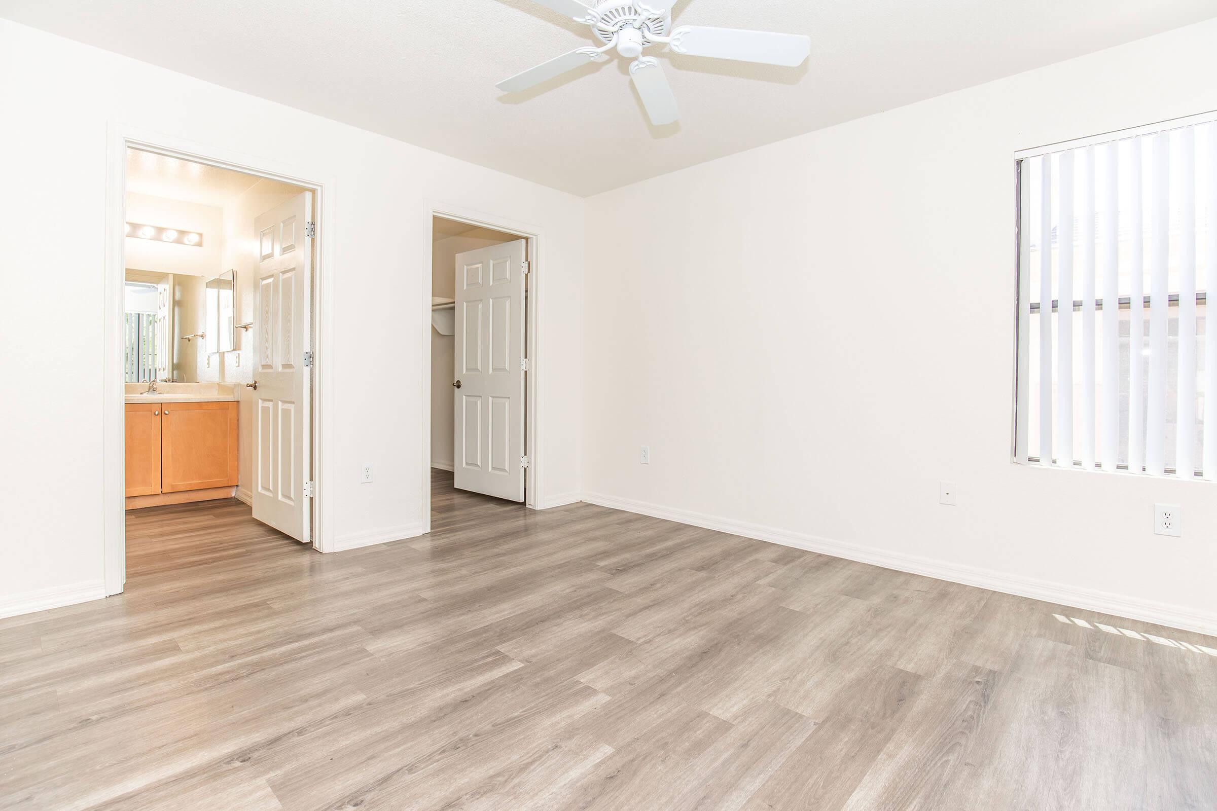 Bright, empty room featuring light-colored walls and a wood-like floor. A ceiling fan is mounted above, and there are two closed doors leading to other spaces. Natural light streams in through a nearby window with blinds. The room has a clean, modern appearance, ready for furniture and personal touches.