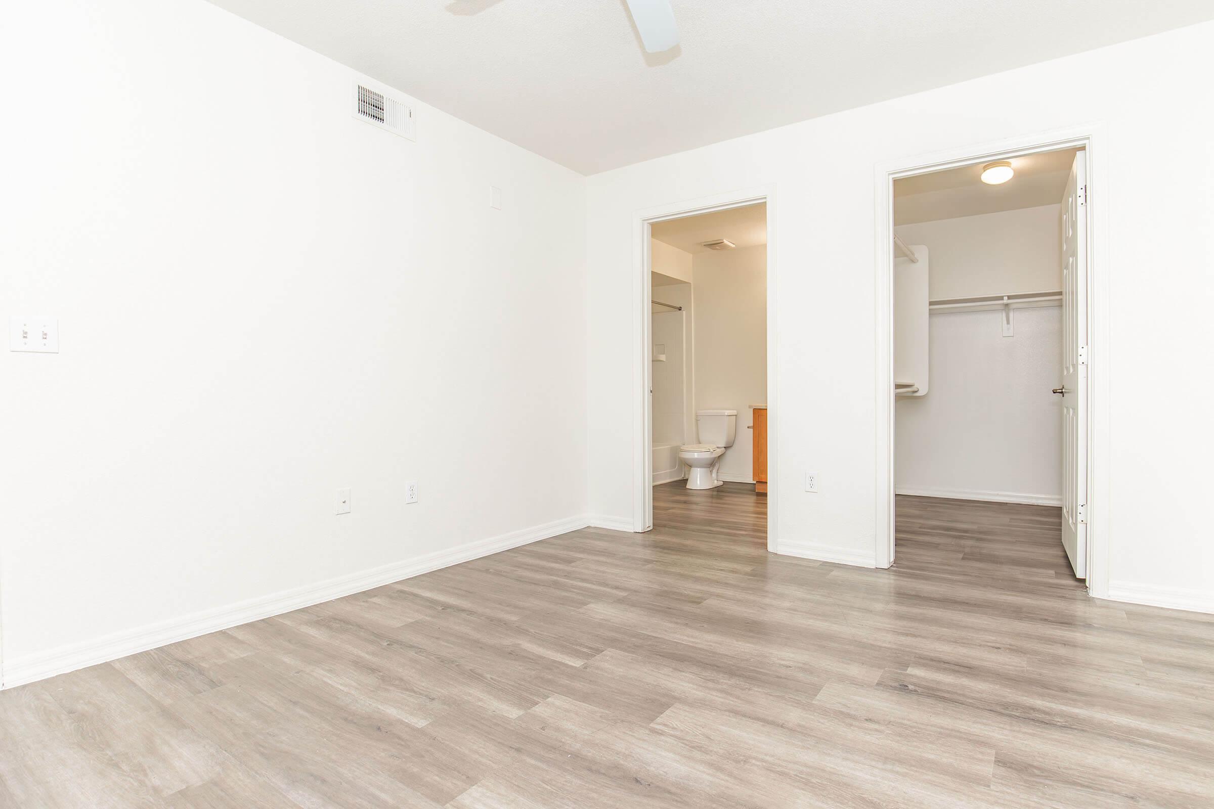 A spacious, empty room with light-colored walls and new laminate flooring. There's a doorway leading to a bathroom on the right, with a visible toilet and a small closet. A ceiling fan hangs above, and the overall atmosphere is bright and clean, suggesting a fresh living space.