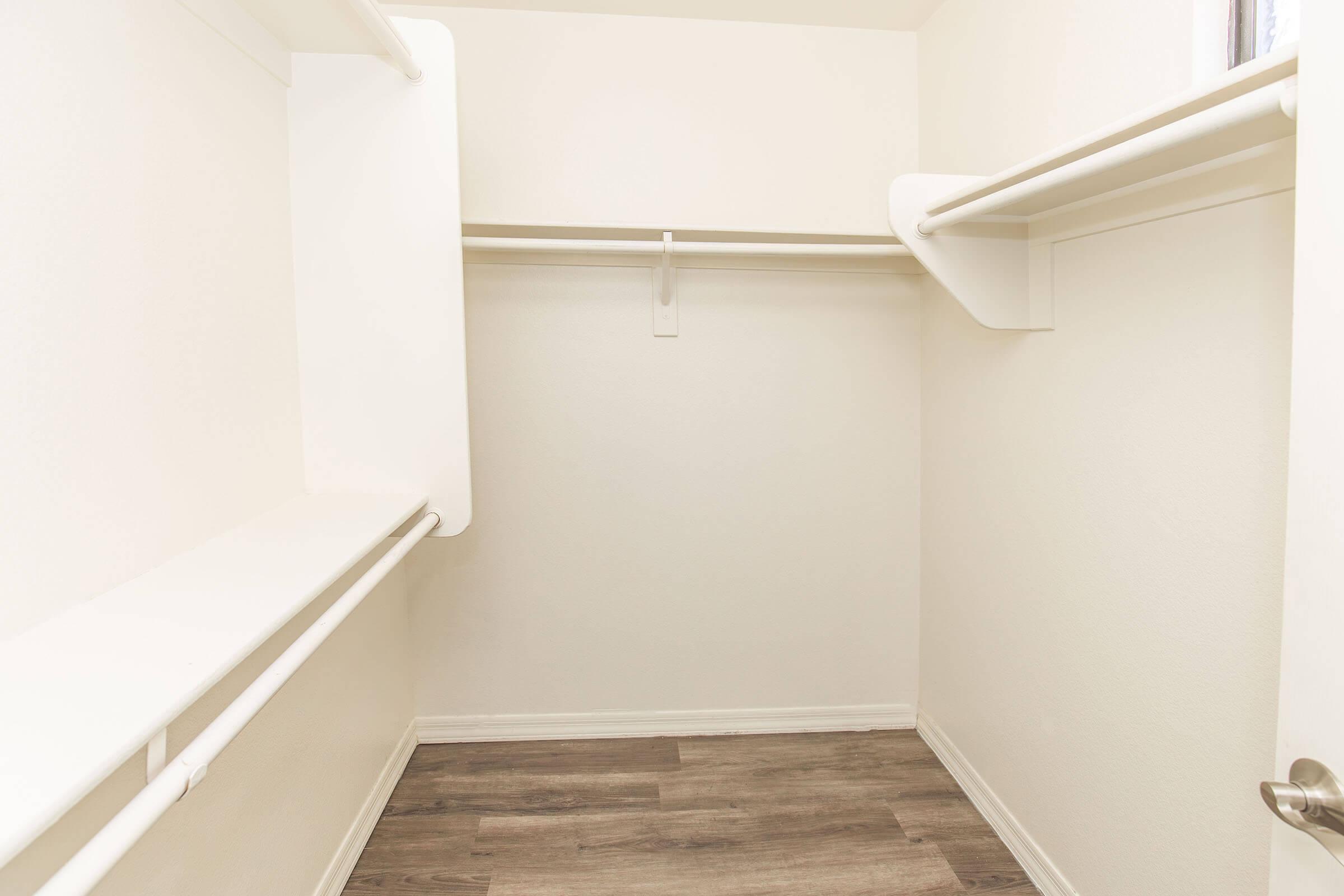 A clean, empty closet with white walls and wooden flooring. There are two horizontal hanging rods along the walls and a shelf above one of the rods, creating a simple and functional storage space. Natural light is coming in from a small window out of view.