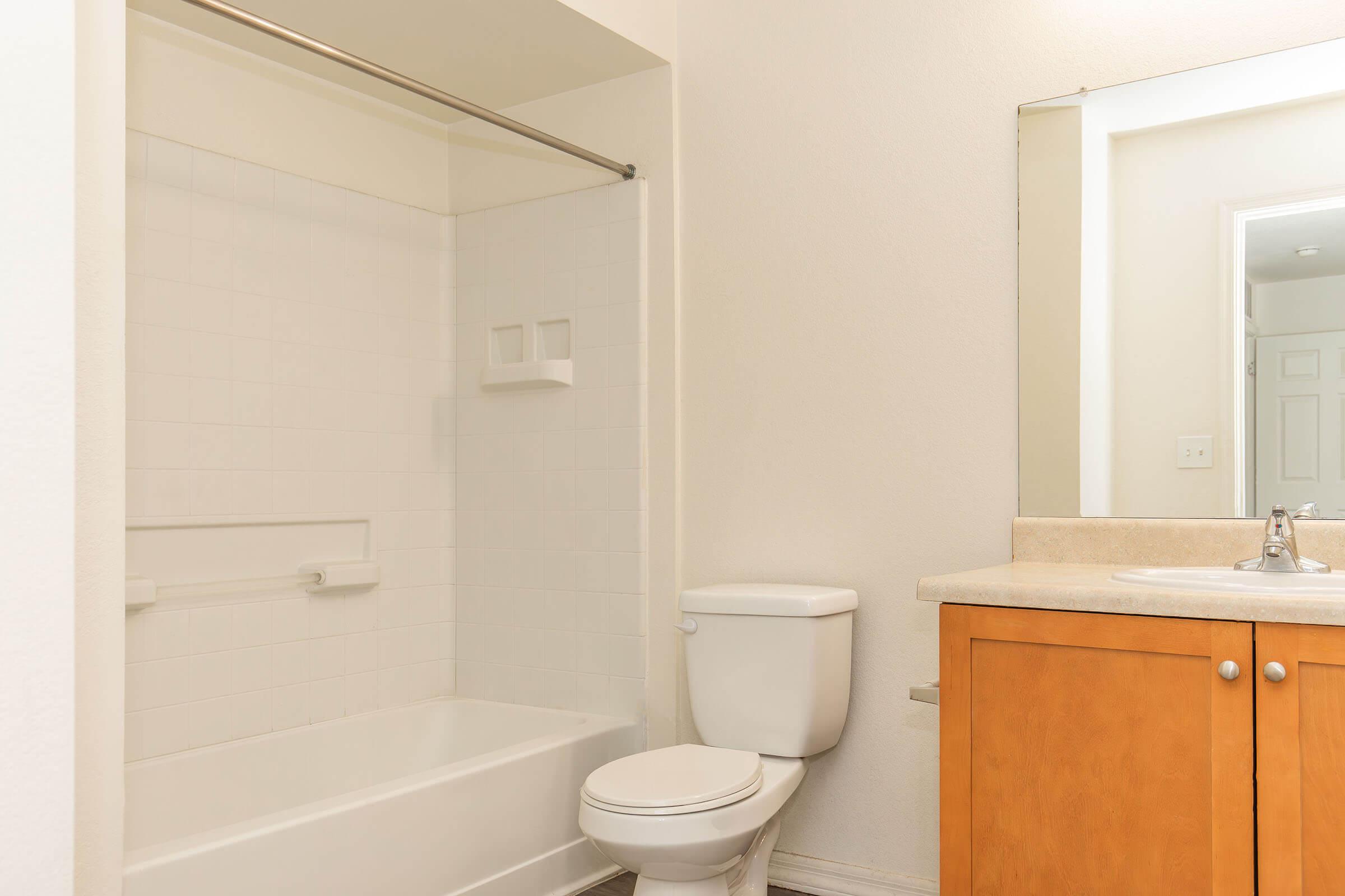 A clean, bright bathroom featuring a white bathtub with a shower curtain rod, a toilet, and a sink with a wooden cabinet underneath. A large mirror is mounted above the sink, reflecting the room's simple, minimalist design with white walls and neutral-colored fixtures.