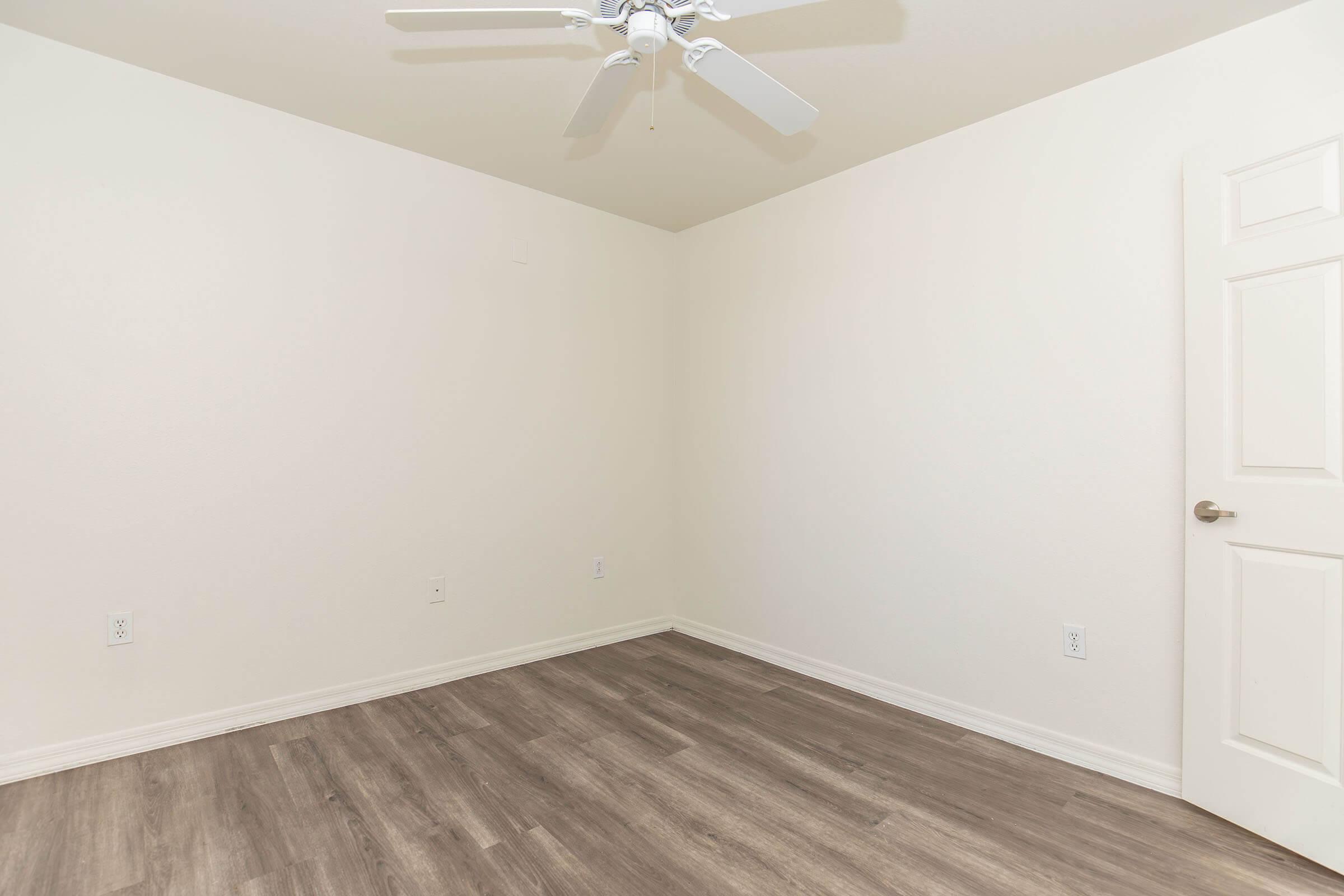 Empty room featuring light-colored walls, a ceiling fan, and a door. The floor is a wood-like laminate. The space is well-lit and spacious, creating a minimalist aesthetic.