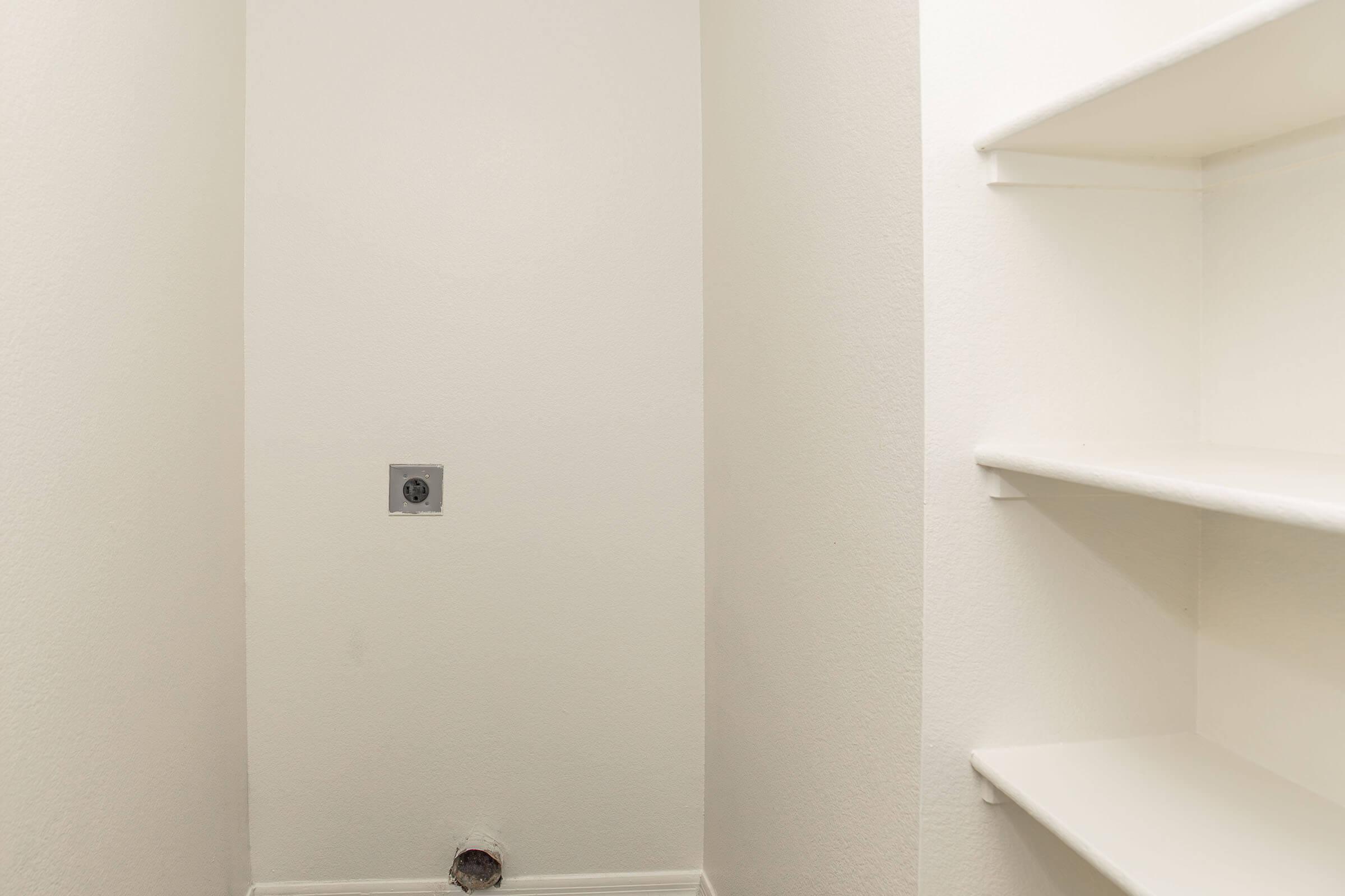 Interior view of a small, empty closet with white walls. There are two shelves on the right side, and a wall-mounted electrical outlet is visible on the left. The floor shows a small vent opening. The space appears clean and well-lit, suitable for storage.