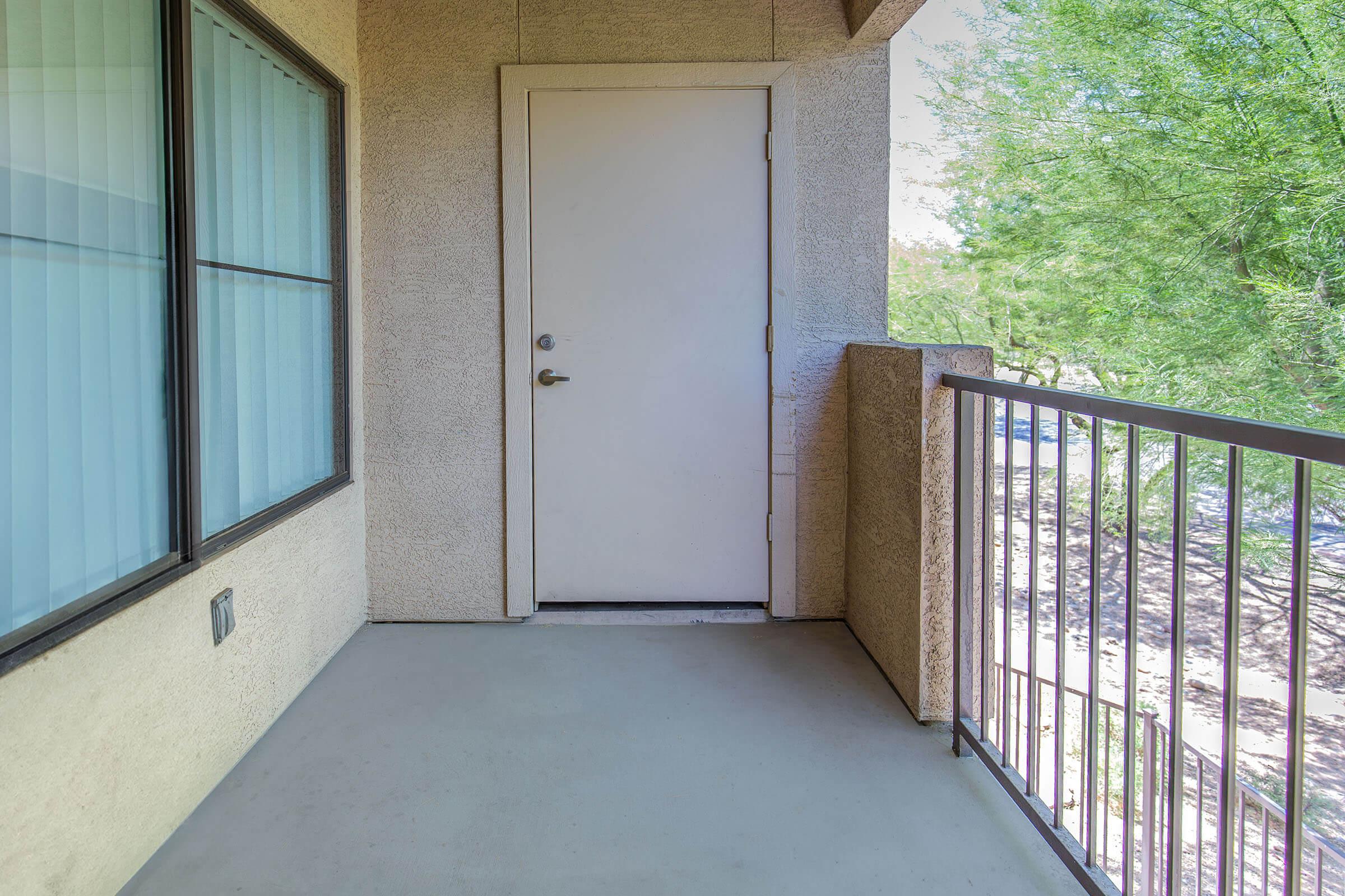 A view of a balcony with a plain door leading inside. The balcony features a railing and is surrounded by greenery visible outside. Natural light illuminates the area, creating a bright and inviting atmosphere.
