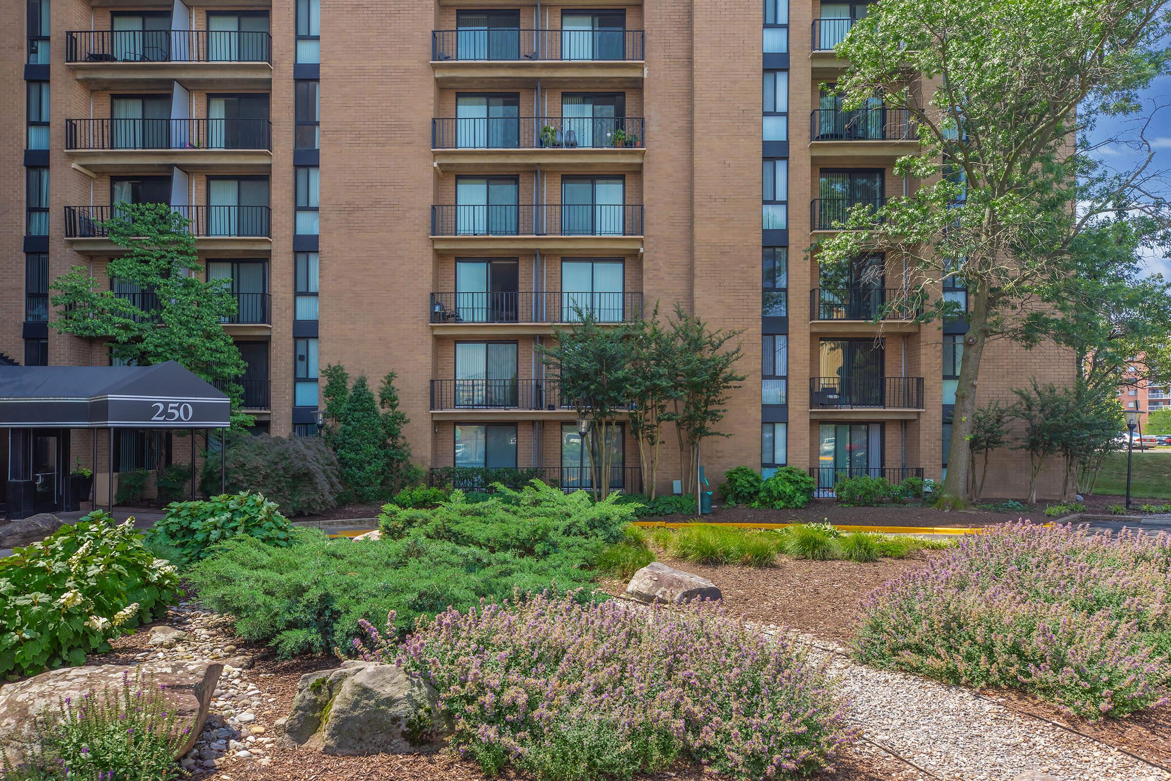 A multi-story brick apartment building with balconies overlooks a landscaped garden featuring low shrubs, flowering plants, and decorative rocks. The building's entrance is marked by a canopy, and there are trees and greenery in the surrounding area. The sky is clear with a few clouds.
