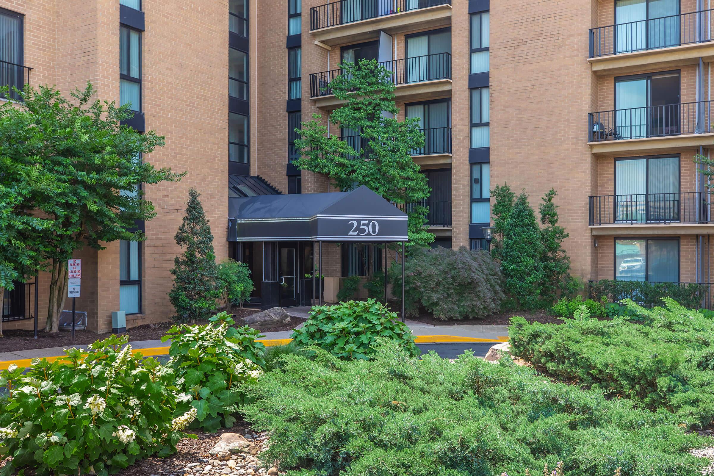 Building facade with a black awning labeled "250," surrounded by landscaped greenery including bushes and trees. There are multiple floors visible, with balconies on some units. The area appears well-kept and is partially sunny.