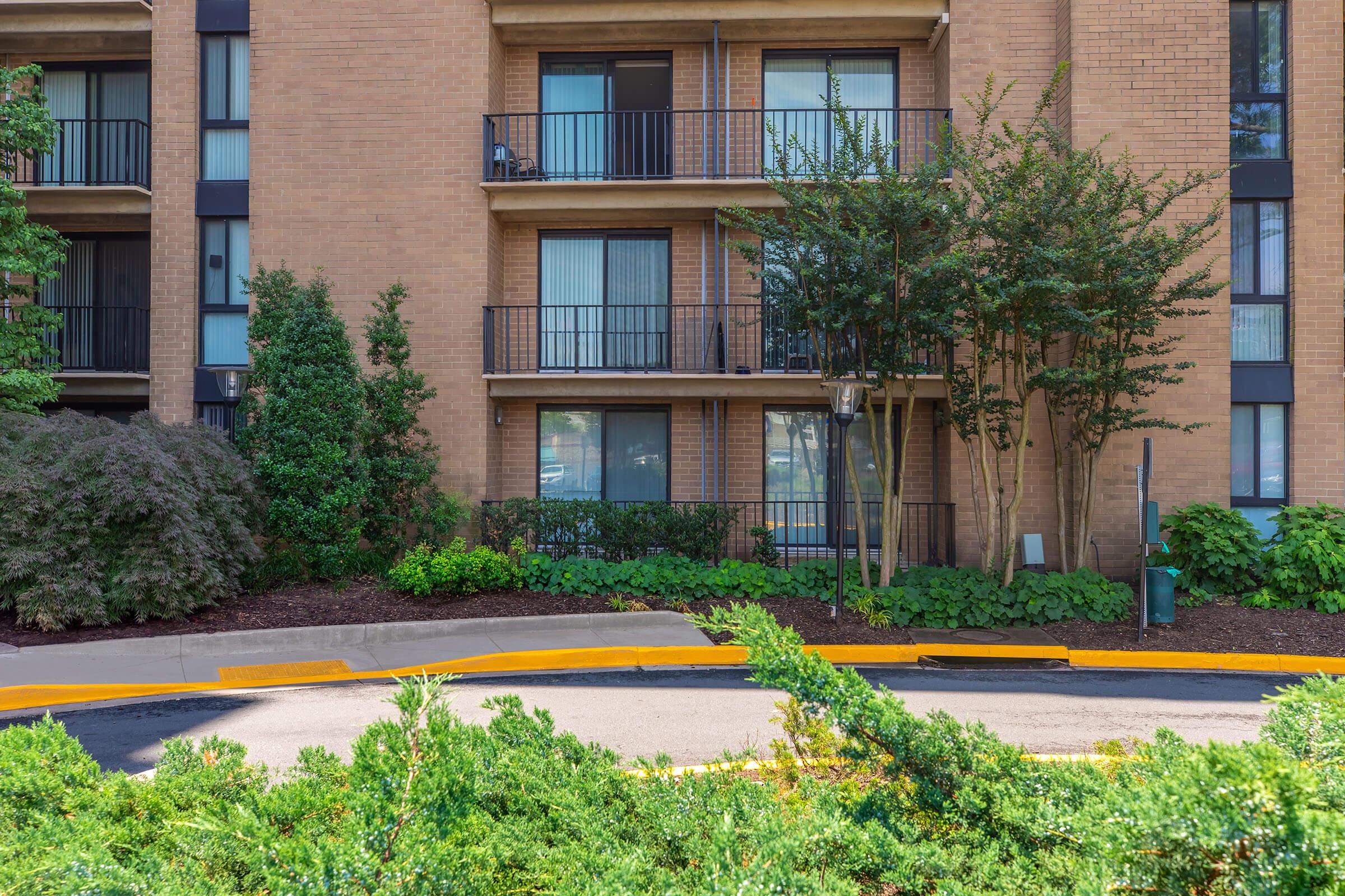 A mid-rise apartment building surrounded by landscaped greenery. The building features balconies and large windows. In the foreground, there's a landscaped area with low shrubs and a curved driveway leading up to the entrance. The setting appears well-maintained and inviting.