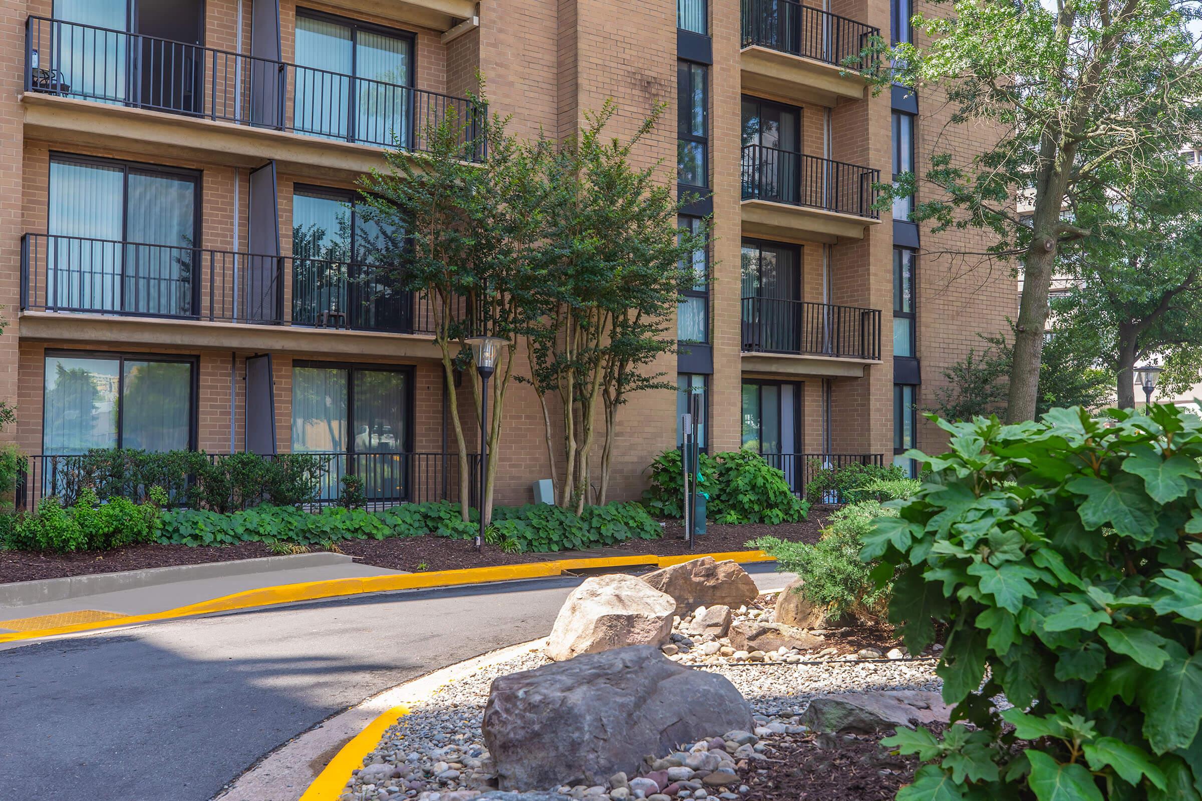 A view of a multi-story residential building featuring balconies, large windows, and landscaped greenery. A winding road leads towards the entrance, with rocks and lush plants along the pathway, creating a welcoming atmosphere.