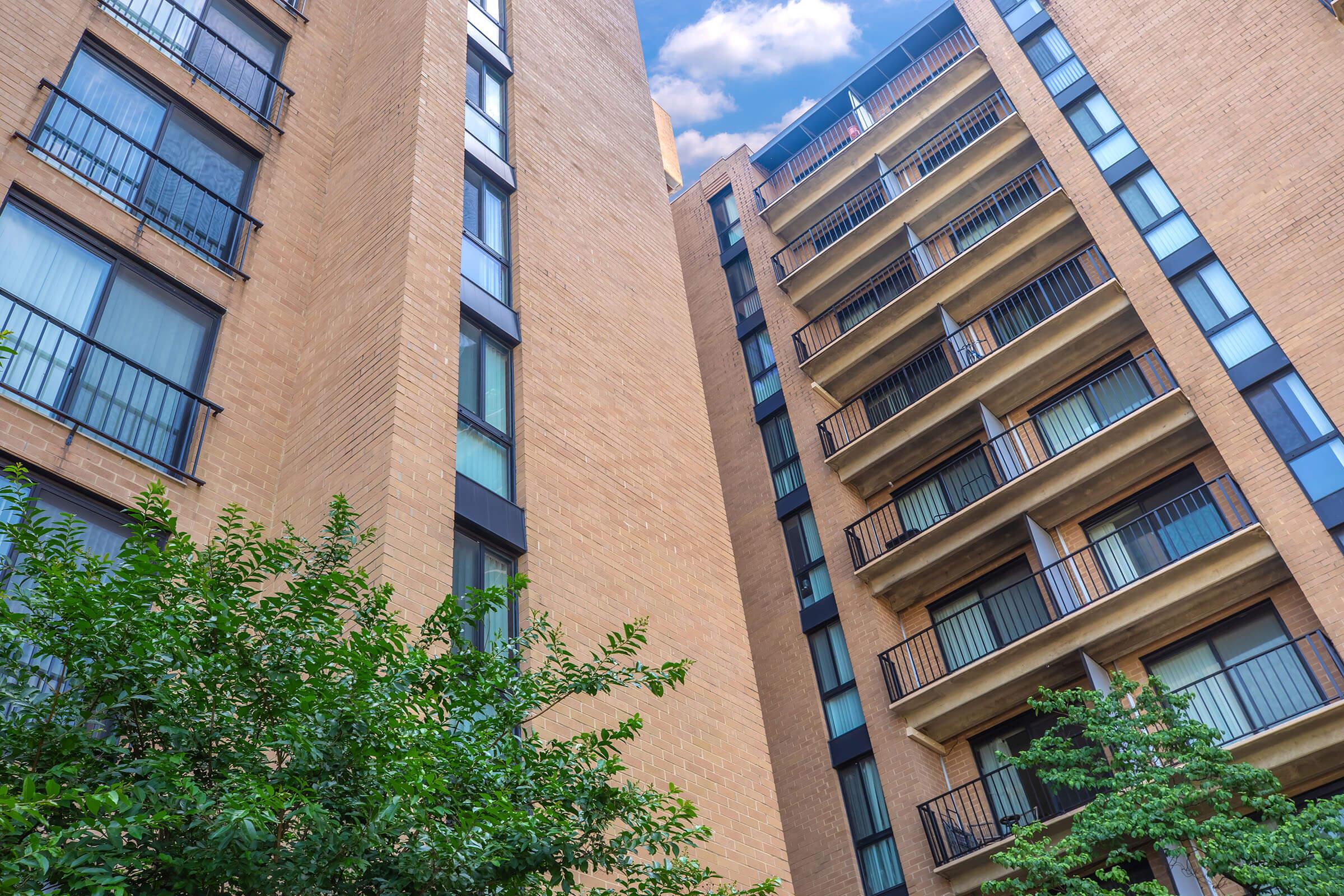 View looking up at a residential building with a brick facade, featuring multiple balconies on the upper floors. Lush greenery is visible in the foreground, contrasting with the building's structure. The sky peeks through the spaces between the buildings, adding depth to the scene.