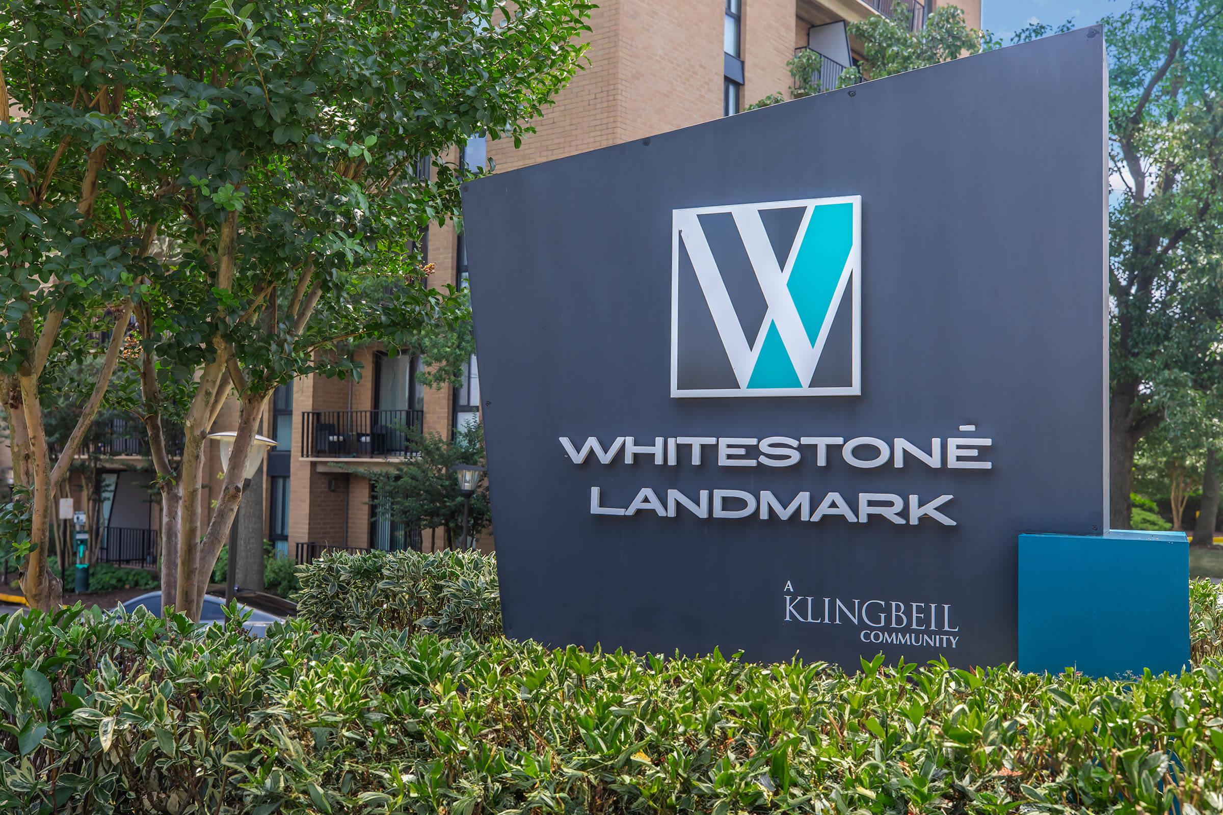 Sign for Whitestone Landmark, a residential community by Klingbeil, featuring a stylized logo with the letter "W" in blue and gray. The sign is surrounded by greenery and located outside a multi-story building.