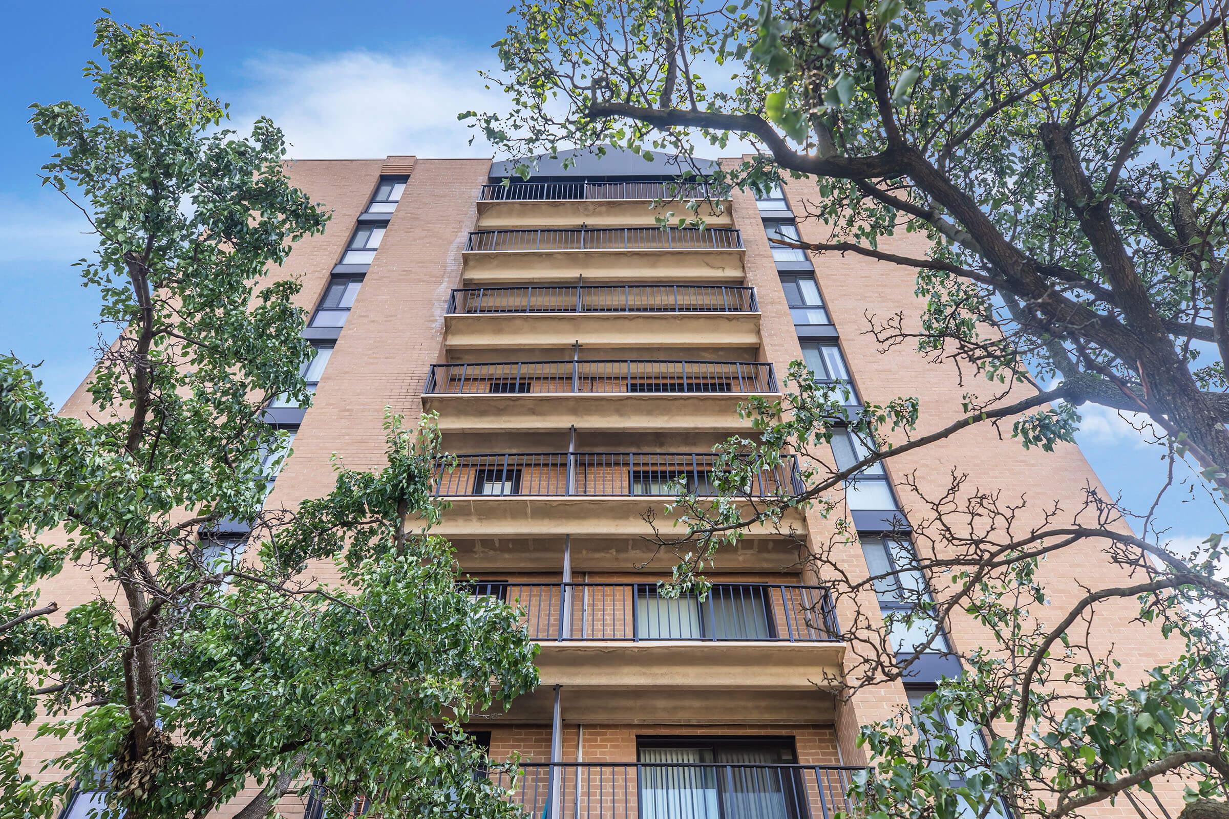 A view of a multi-story brick building featuring several balconies, framed by trees in the foreground. The sky is partly cloudy, and sunlight is visible on the building's facade, highlighting its architectural details.