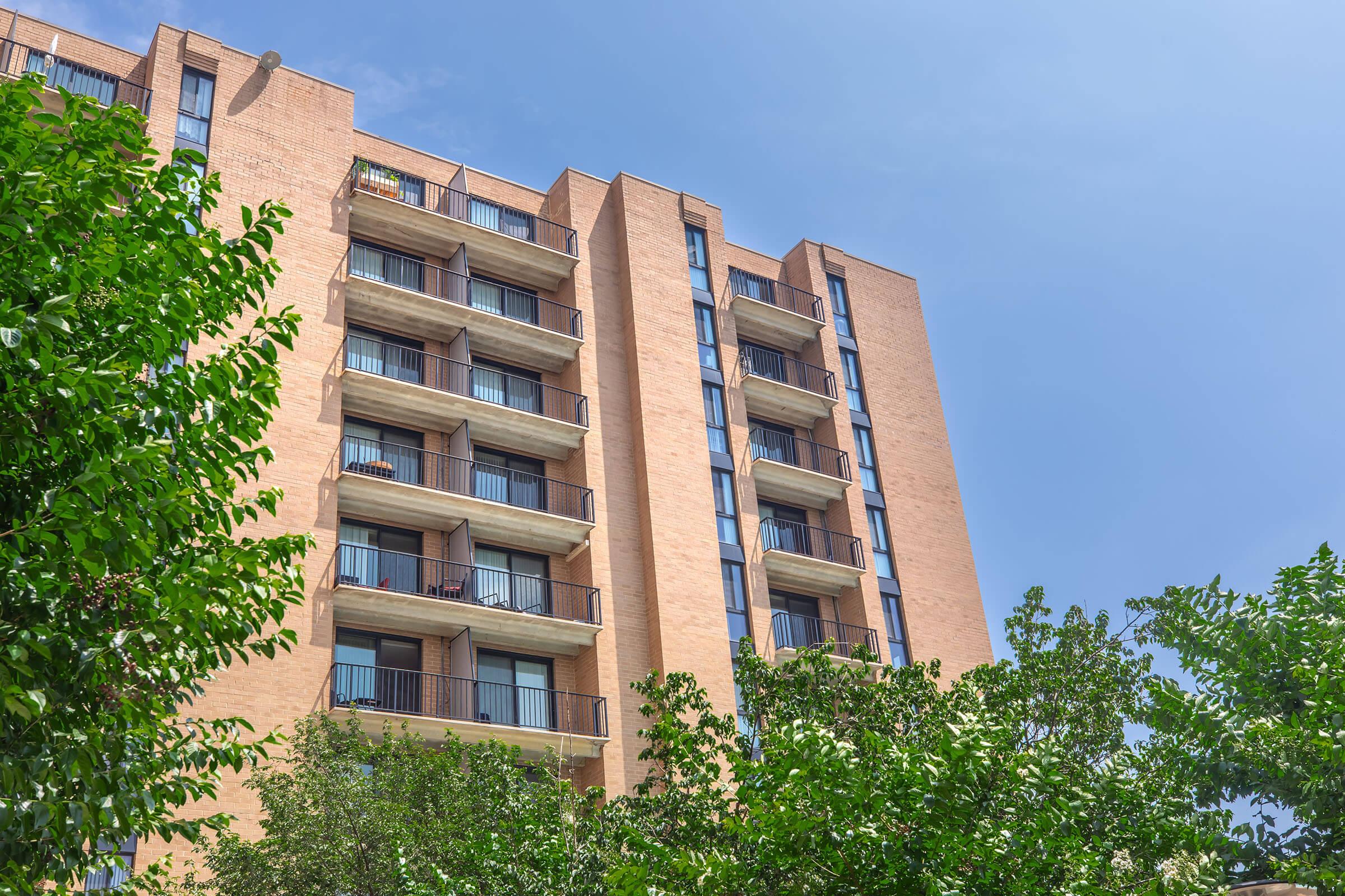 A multi-story brick apartment building with balconies on each floor, surrounded by green trees. The sky is clear with a few clouds, creating a bright and inviting atmosphere.