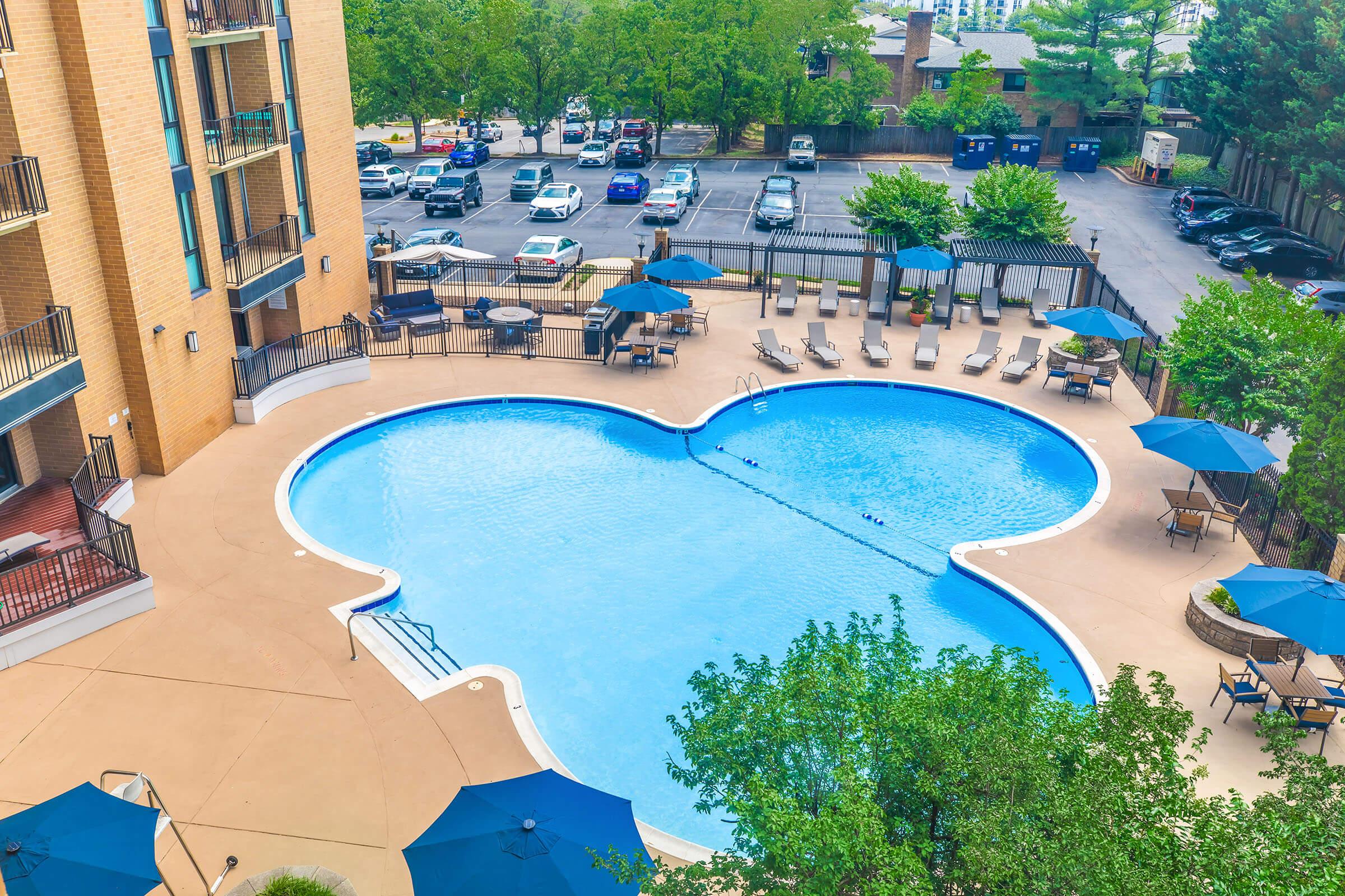A view of a swimming pool shaped like a Mickey Mouse head, surrounded by lounge chairs and umbrellas. Green trees and a parking lot with several cars are visible in the background. The setting appears to be part of a residential or leisure complex.