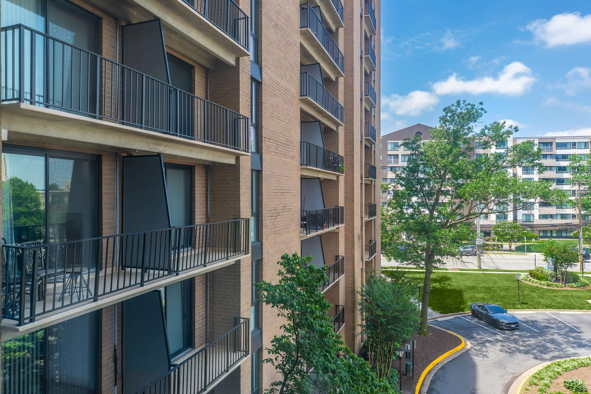 View of a multi-story apartment building with balconies, featuring modern architecture. There are green trees and landscaped areas in the foreground, and a small vehicle parked on a circular driveway. The sky is blue with scattered clouds, creating a bright and inviting atmosphere.