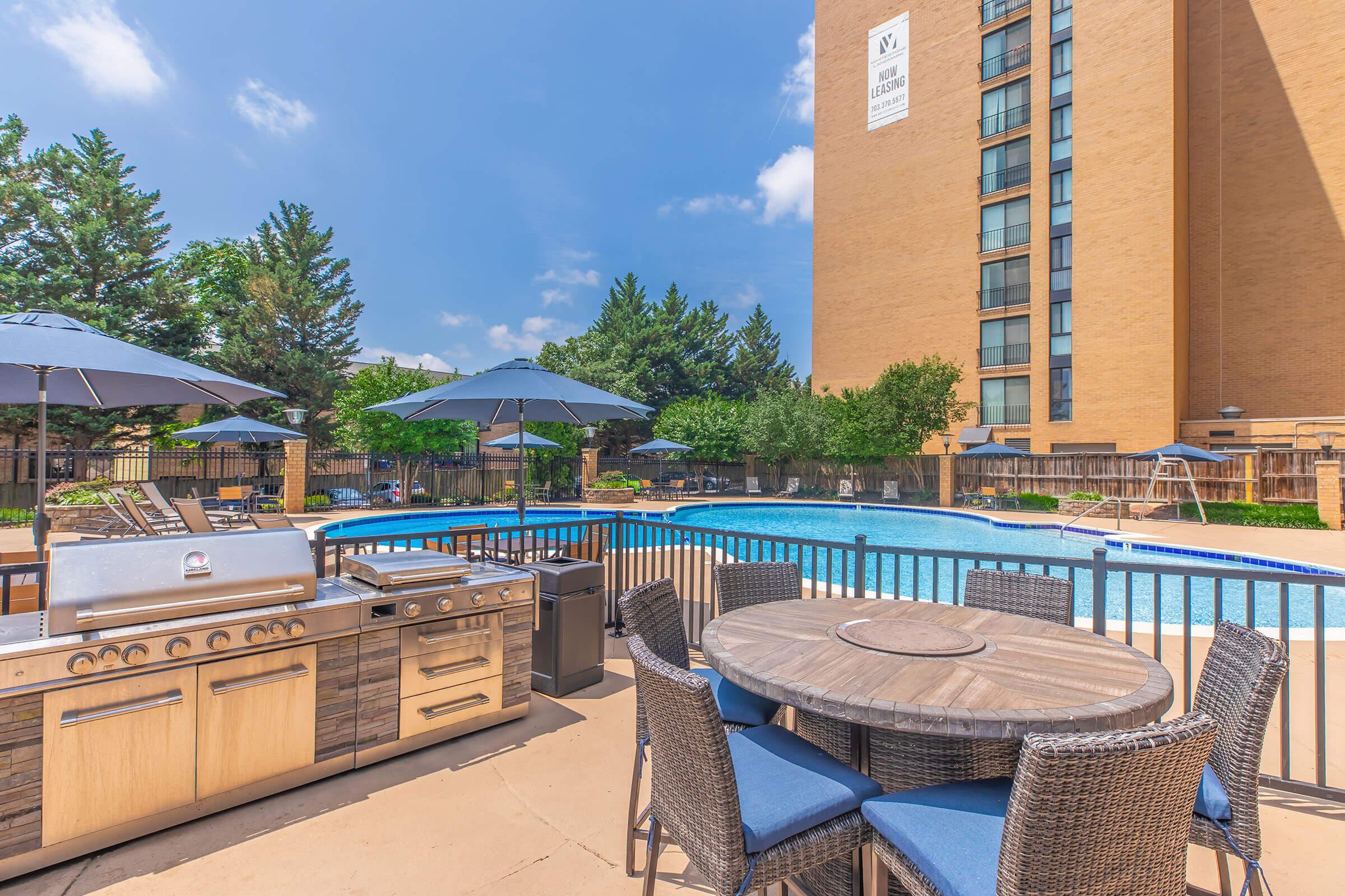 Outdoor area featuring a swimming pool surrounded by greenery, with a barbecue grill and a dining table set near the pool. Umbrellas provide shade over the seating area, and a tall building is visible in the background under a clear blue sky.