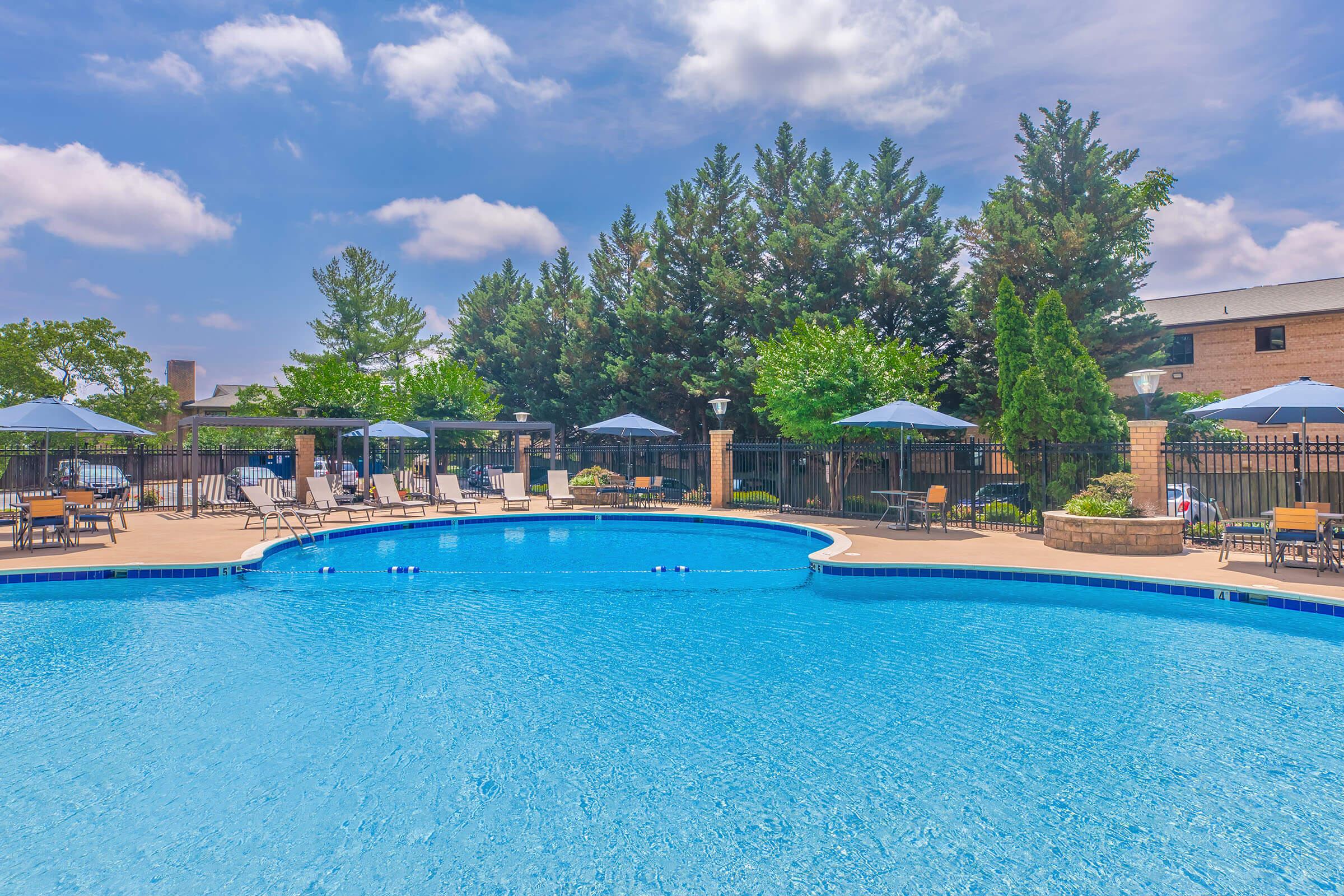 A large swimming pool surrounded by lounge chairs and umbrellas, set against a backdrop of trees and a clear blue sky. The pool features a gentle curve, with clear water reflecting the sunny day. Nearby, there are shaded seating areas and a fence enclosing the pool area.