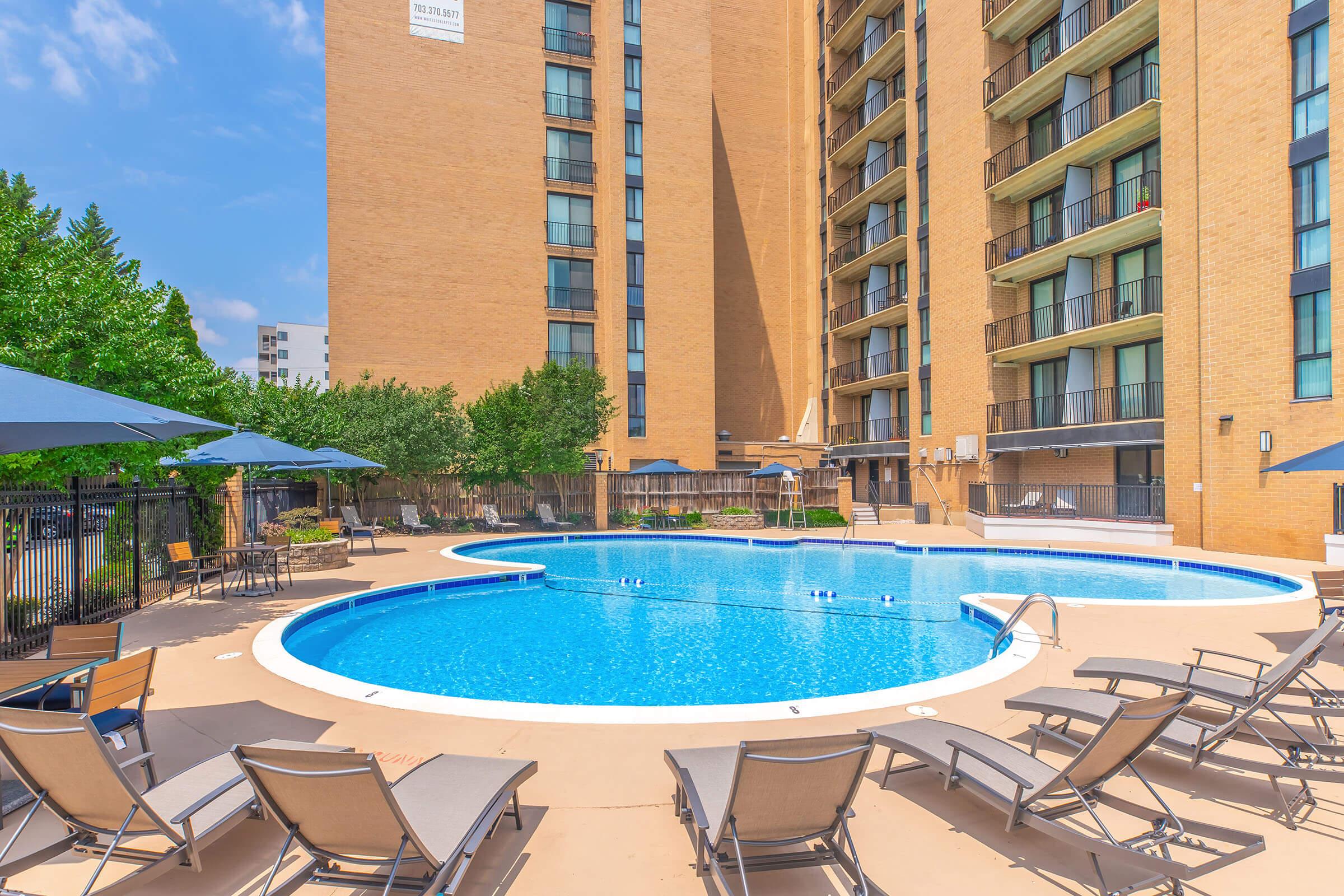 A spacious outdoor pool surrounded by lounge chairs and umbrellas, located in a residential area. The backdrop features a tall, light-colored building with balconies and greenery around the pool area, creating a relaxing and inviting atmosphere on a sunny day.