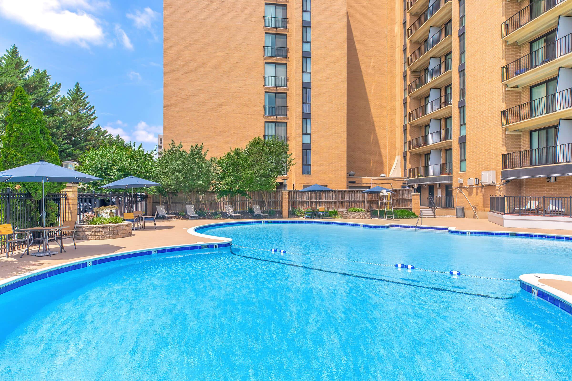 A large outdoor swimming pool surrounded by lounge chairs and shaded umbrellas, with a brick building in the background. Lush greenery adds to the relaxing atmosphere on a sunny day, providing a tranquil setting for leisure and relaxation.