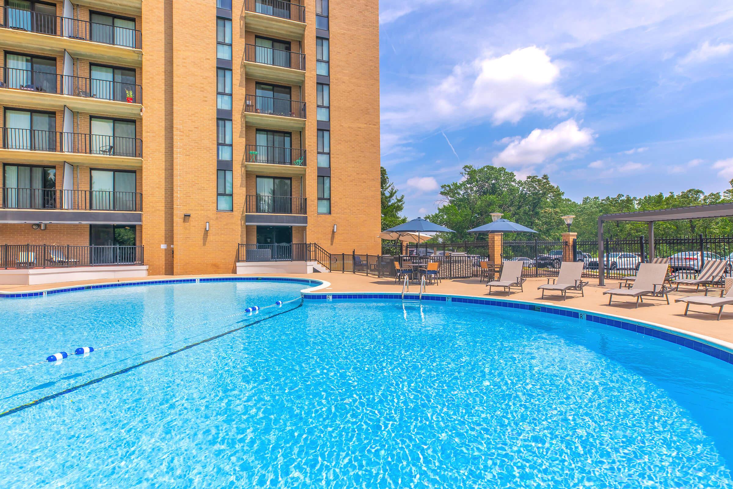 A clear blue swimming pool with lounge chairs surrounding it, located beside a multi-story brick building. The pool area features umbrellas for shade, and the sky above is bright with a few clouds. Green trees are visible in the background, adding to the tranquil atmosphere.