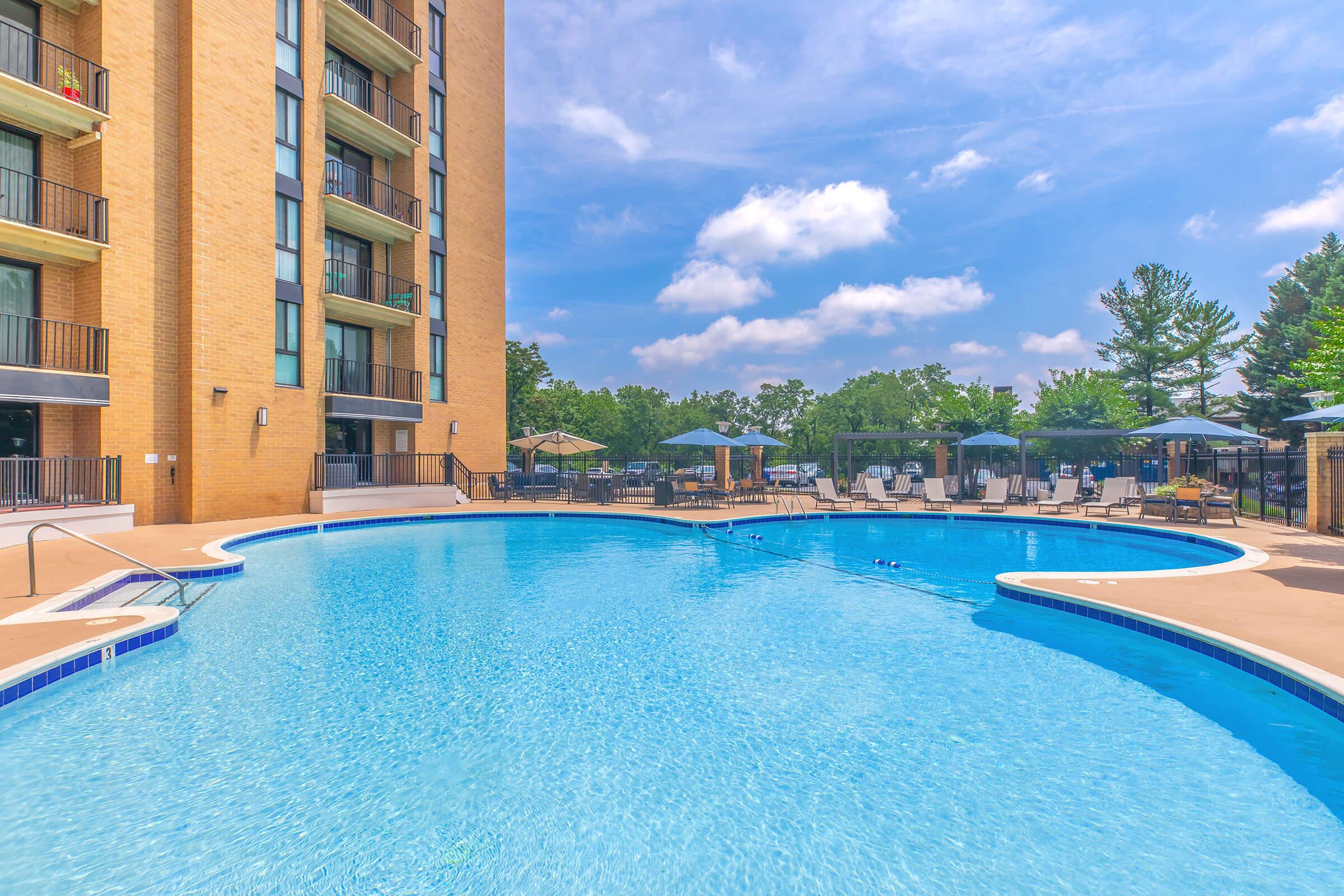 A large swimming pool with clear blue water, surrounded by lounge chairs and umbrellas. In the background, a tall brick building can be seen, along with green trees and a bright blue sky with fluffy clouds. The setting appears to be a resort or a residential complex.