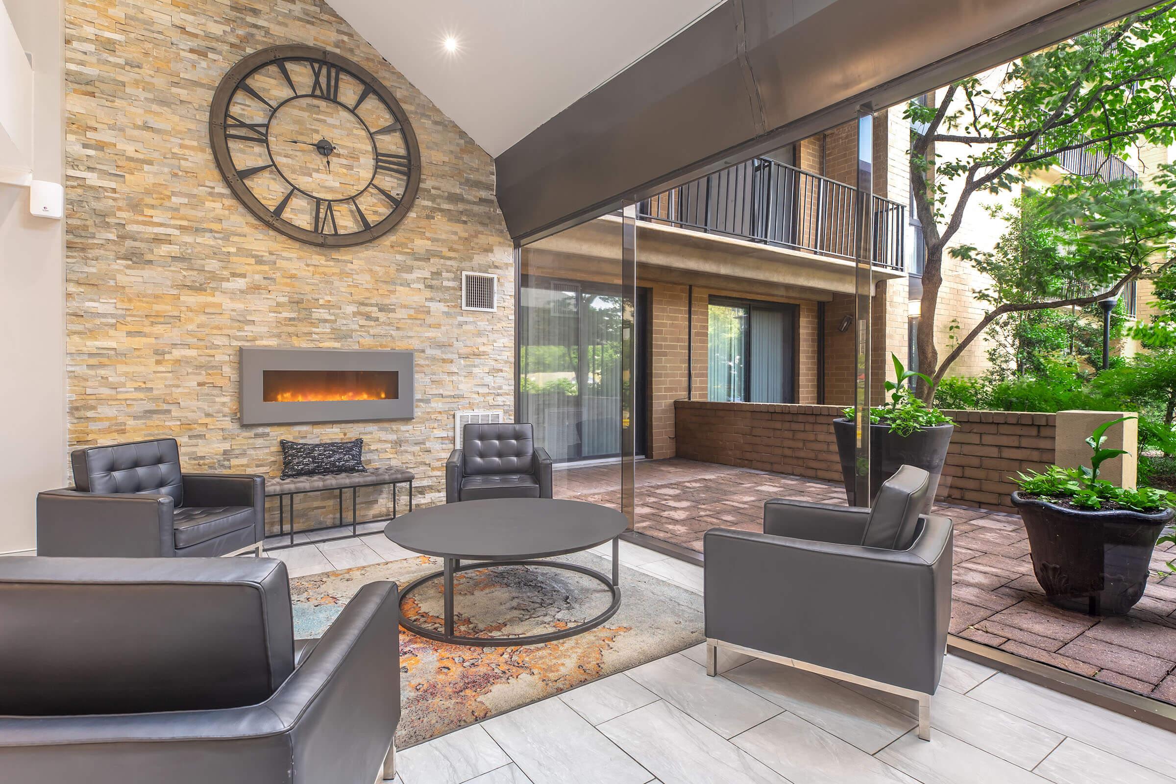 A modern lounge area featuring a stone accent wall with a large clock, a built-in fireplace, and contemporary furniture including black leather chairs and a round table. Large glass windows offer a view of an outdoor patio with greenery, creating a bright and inviting atmosphere.