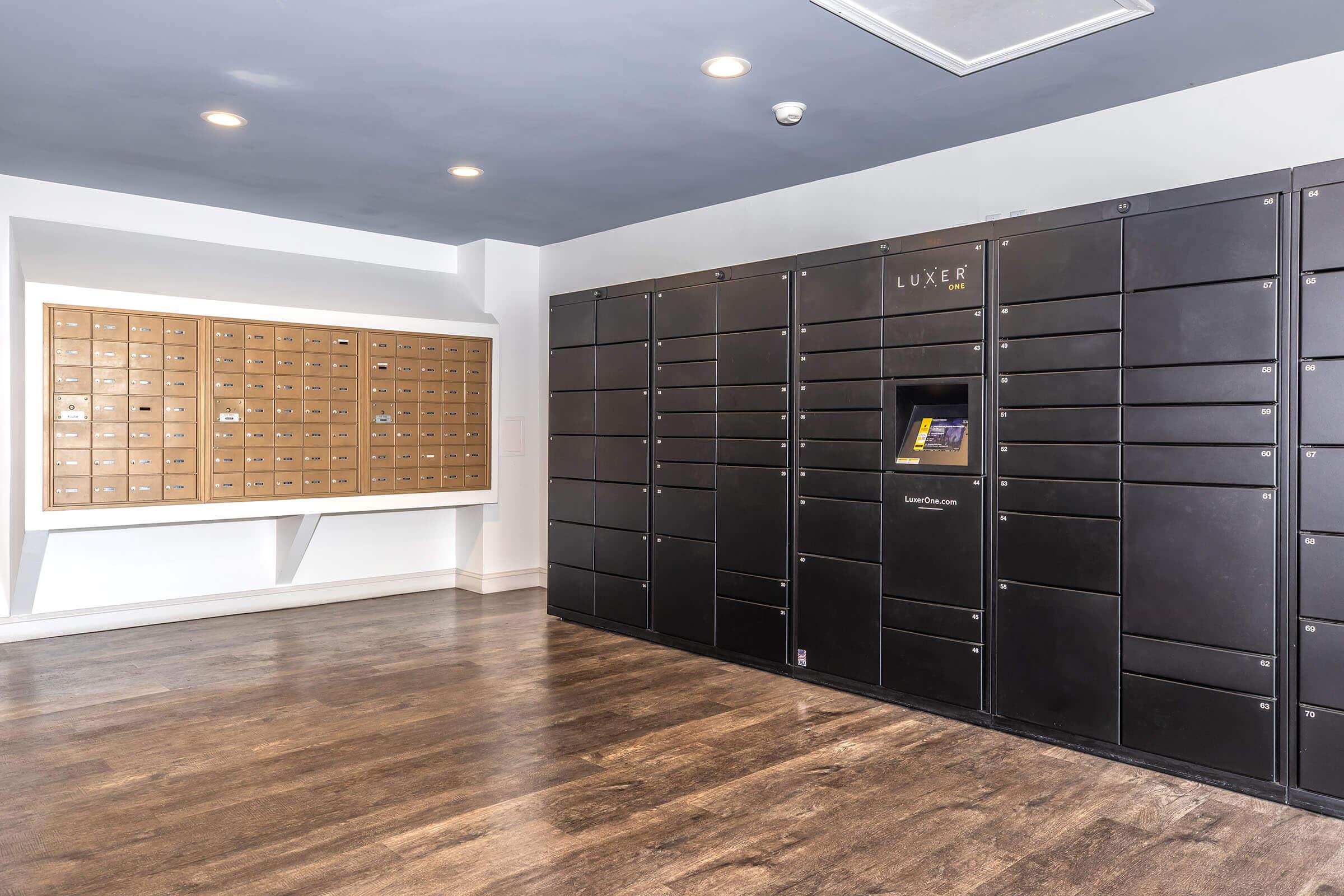 Interior view of a modern mailroom featuring two sets of mail lockers. On the left, a wall-mounted set with numerous small compartments, and on the right, a sleek black locker system with a touchscreen interface for package retrieval. The flooring is wooden, and the lighting is bright.