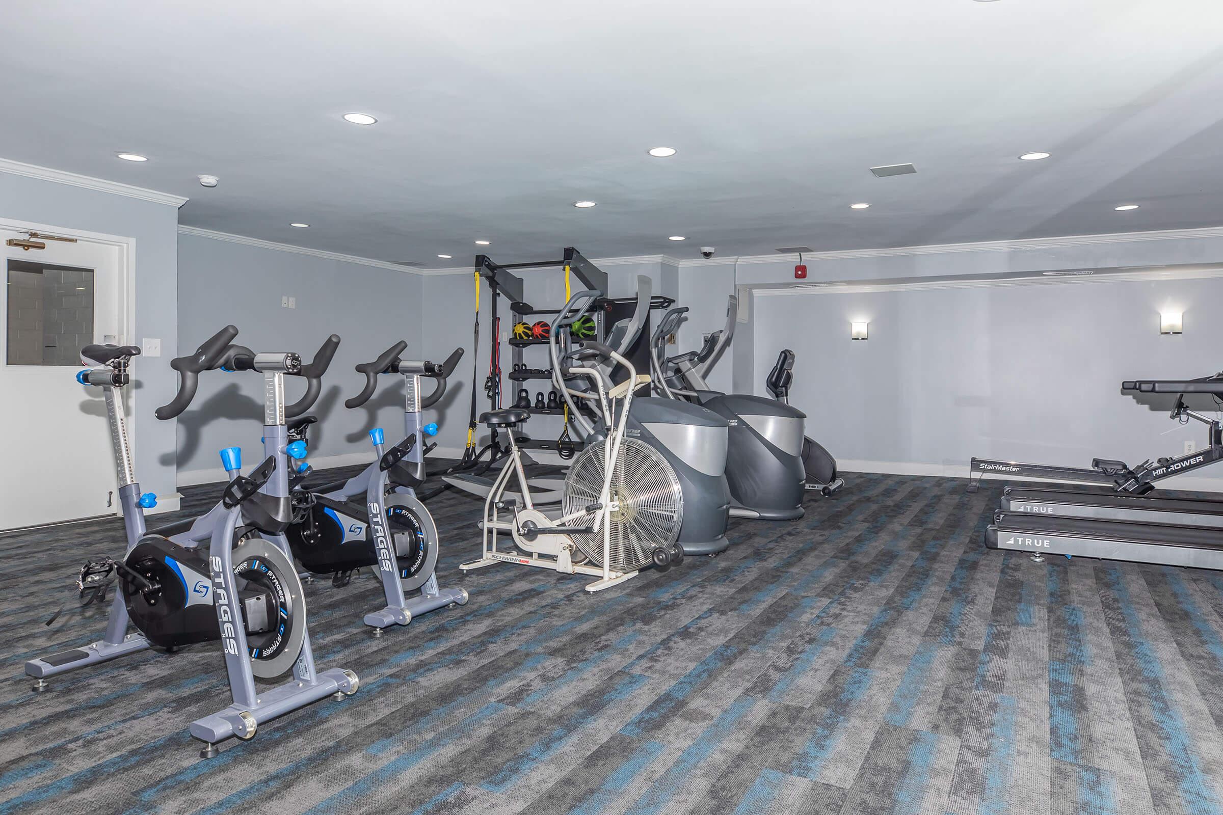 A modern gym featuring various exercise equipment including stationary bikes, elliptical machines, a rowing machine, and free weights. The space has a light gray and blue color scheme with carpeting and wall-mounted lighting. The room appears well-lit and organized for fitness activities.