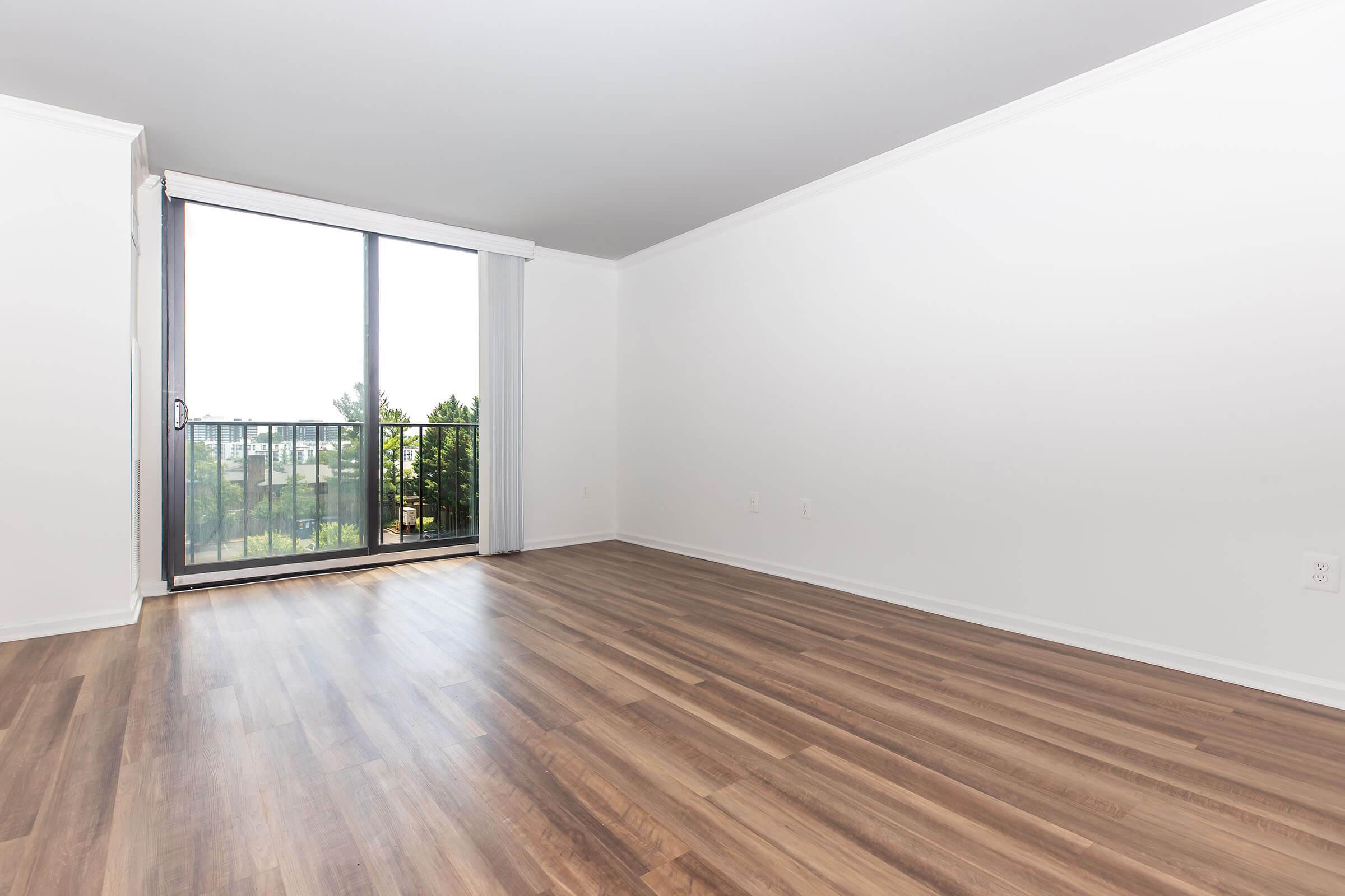 Empty room with light-colored walls and modern wood flooring. A large window with a sliding glass door offers a view of greenery outside. The room is bright and spacious, providing a serene atmosphere. Natural light fills the space, enhancing its openness.