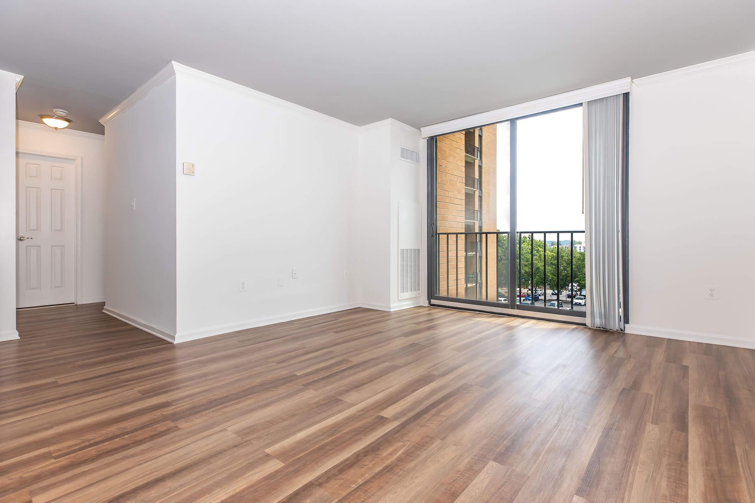 Bright and spacious interior of a modern apartment featuring light-colored walls, a large window with a balcony view, and stylish wooden flooring. The room is empty, creating a sense of openness and potential for personalization. Light fixtures and a door to another room are visible.