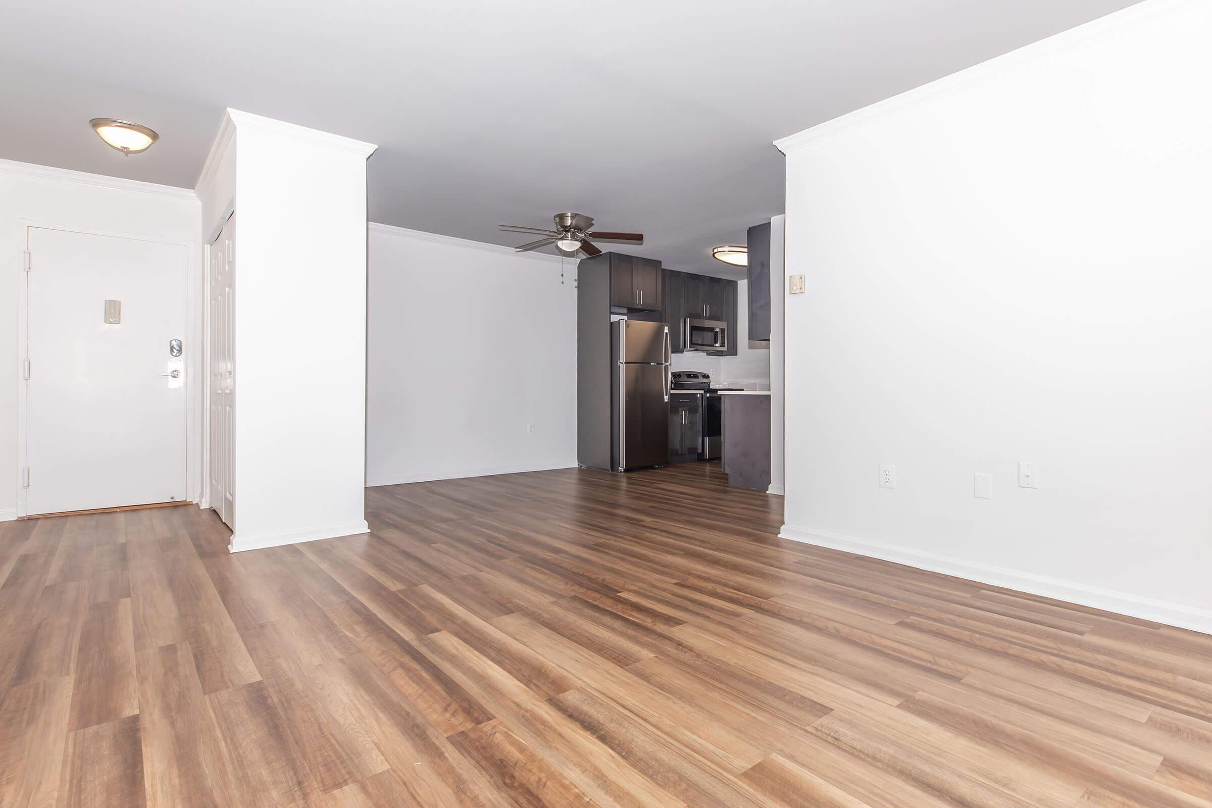 A spacious, well-lit room with wooden flooring, featuring a ceiling fan. In the background, there is a modern kitchen with stainless steel appliances and cabinetry. The walls are painted white, contributing to a clean and open atmosphere, ideal for a living space or apartment setting.