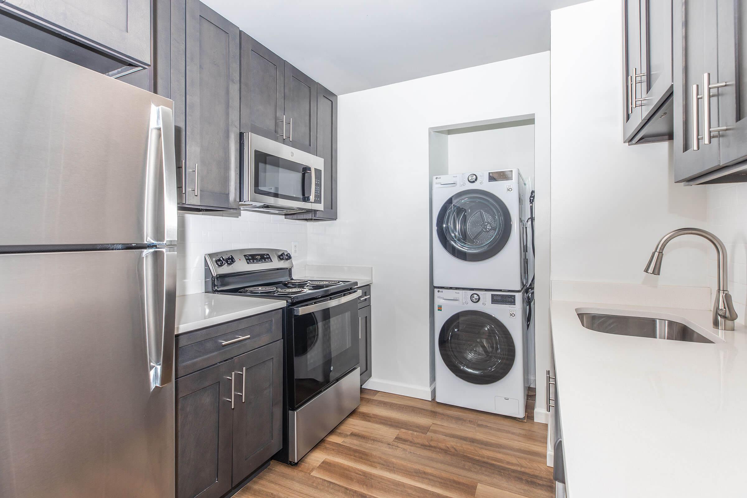 A modern kitchen featuring stainless steel appliances, including a refrigerator, microwave, and oven. There's a stacked washer and dryer in a recessed area. The cabinetry is dark wood, and the countertops are light. The flooring is wooden, creating a warm and contemporary ambiance.