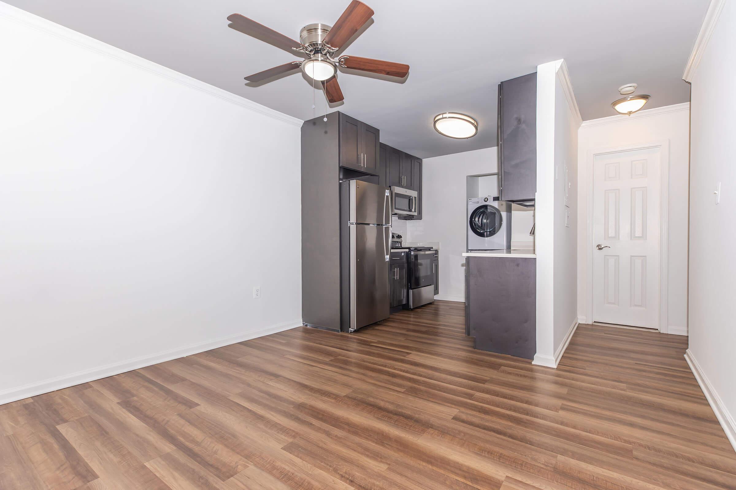 A modern kitchen and living space featuring brown hardwood flooring, a ceiling fan, stainless steel appliances, a washer dryer unit, and minimalist cabinetry. The walls are painted white, creating a bright and open atmosphere.
