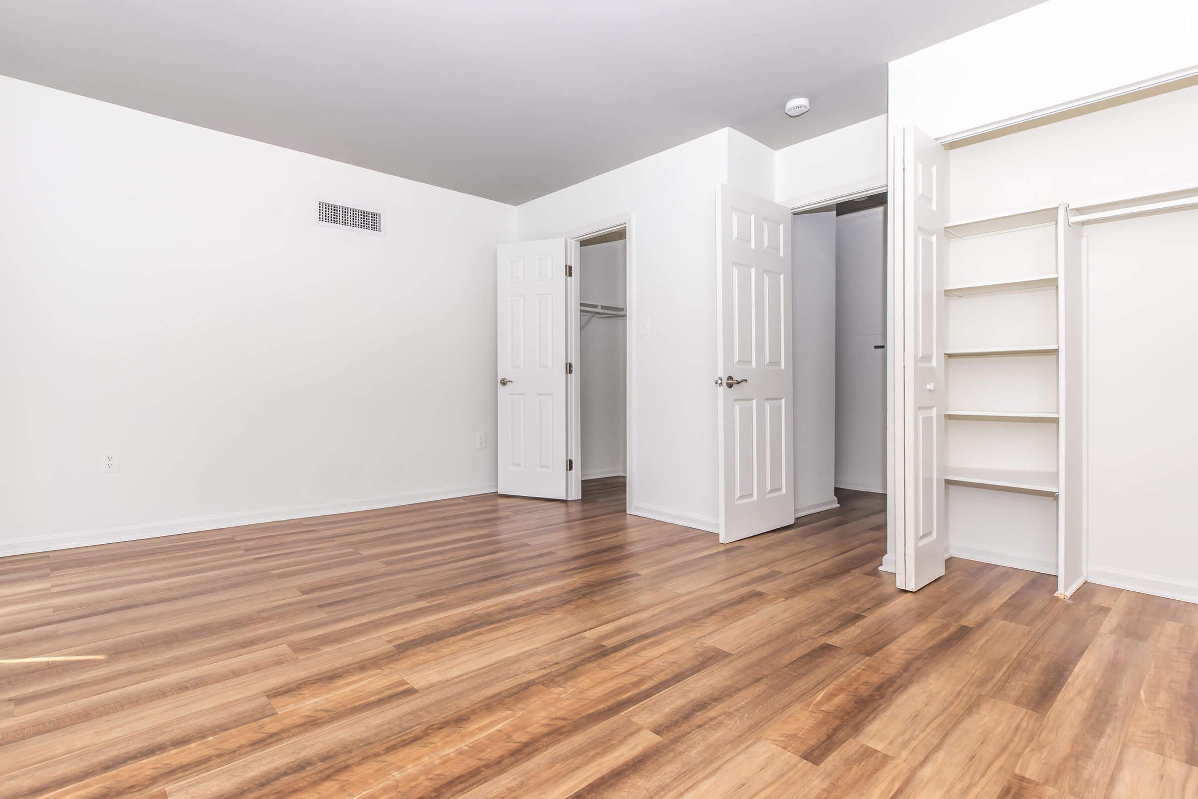 A spacious, empty room with light-colored walls and wooden flooring. Two open closet doors reveal additional storage space, and a third door leads to another area. Natural light fills the room, creating a bright and airy atmosphere.