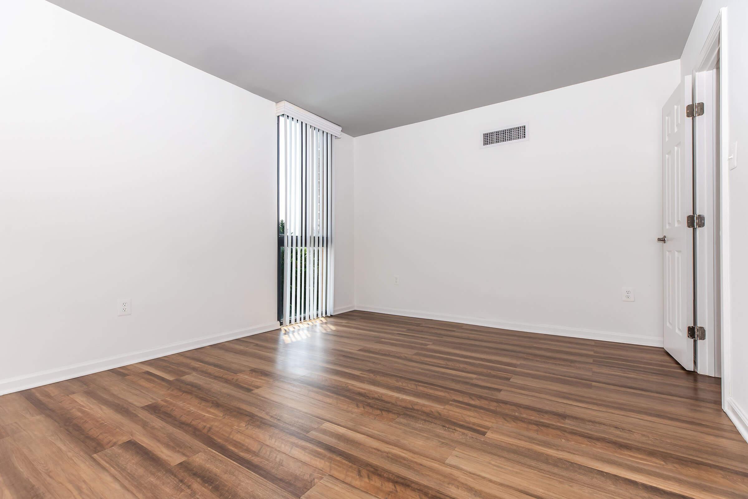 A spacious, empty room with light hardwood flooring. The walls are painted white, and there are vertical blinds covering a window on the left side. A door is visible on the right, leading to another area. The room is well-lit, and the absence of furniture emphasizes its openness.