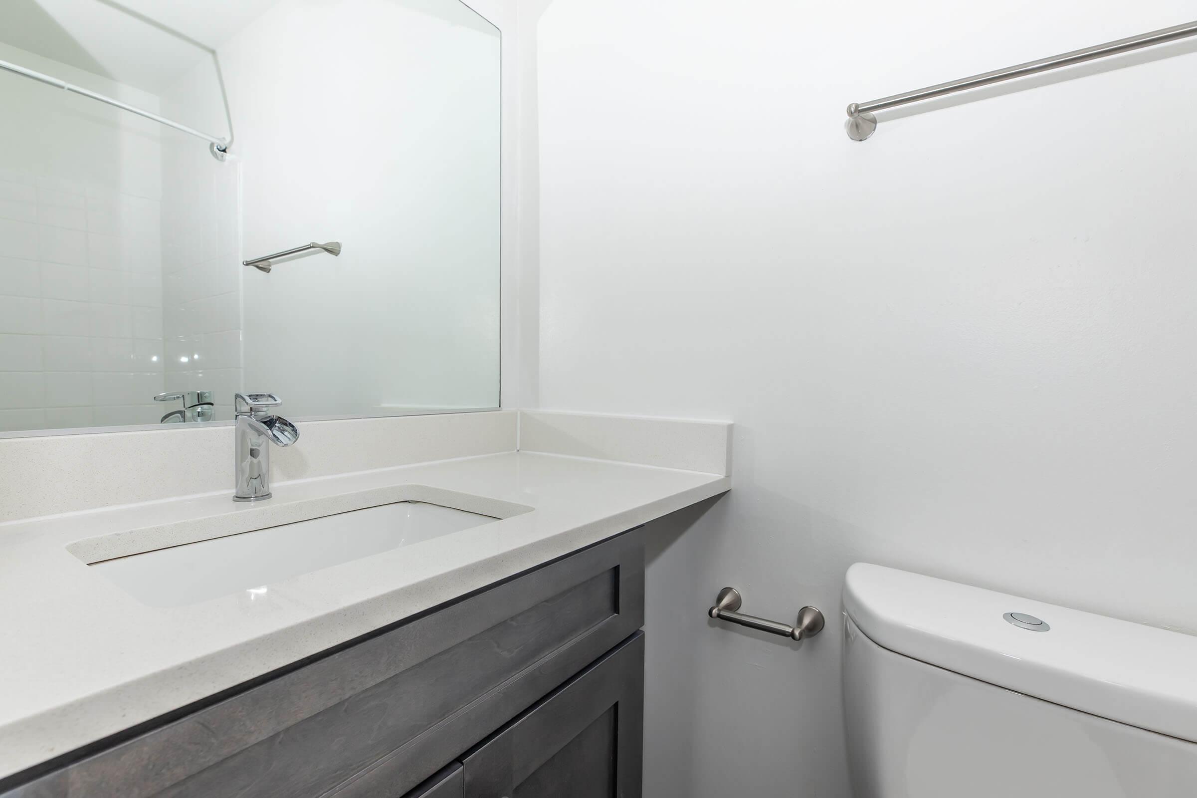 A modern bathroom featuring a sleek sink with a faucet mounted on a light-colored countertop. A large mirror is above the sink, and there's a toilet to the right. The walls are painted white, and a towel bar is mounted beside the mirror. The overall design is minimalistic and clean.