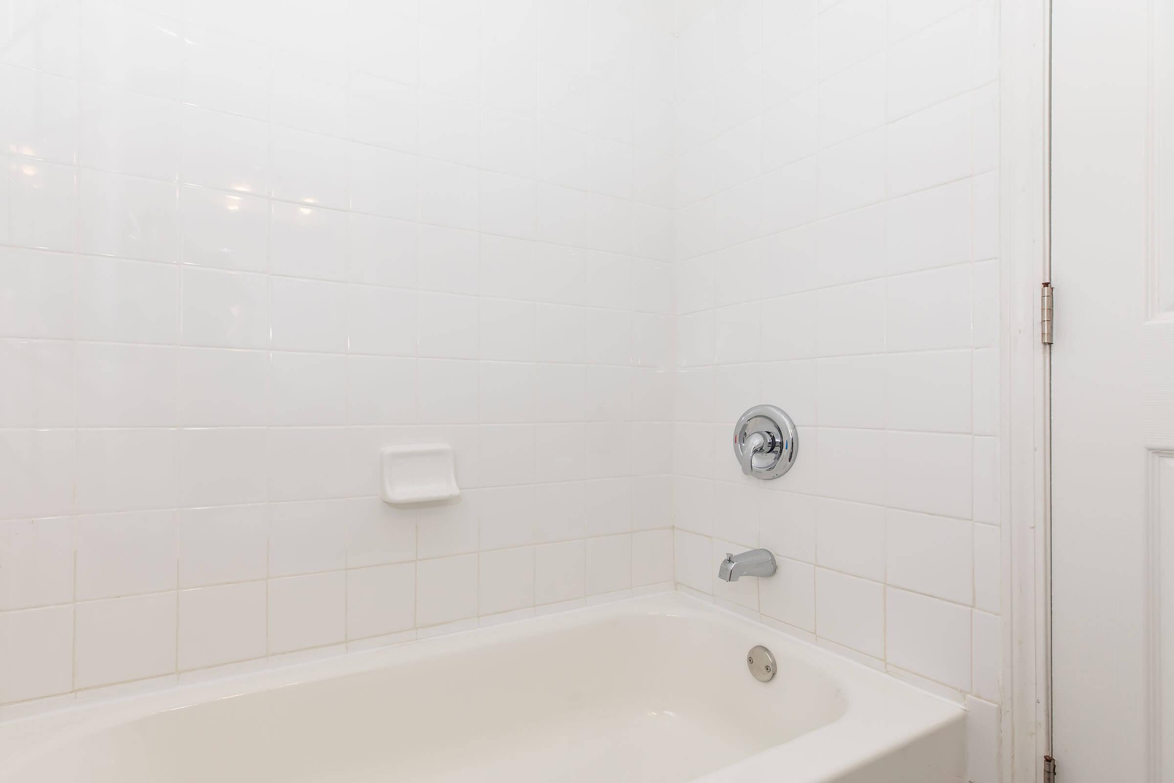 White tiled bathroom with a bathtub on the left side and a faucet on the wall. There is a grab bar for safety and a soap dish mounted on the wall. The lighting is bright, creating a clean and minimalist appearance.