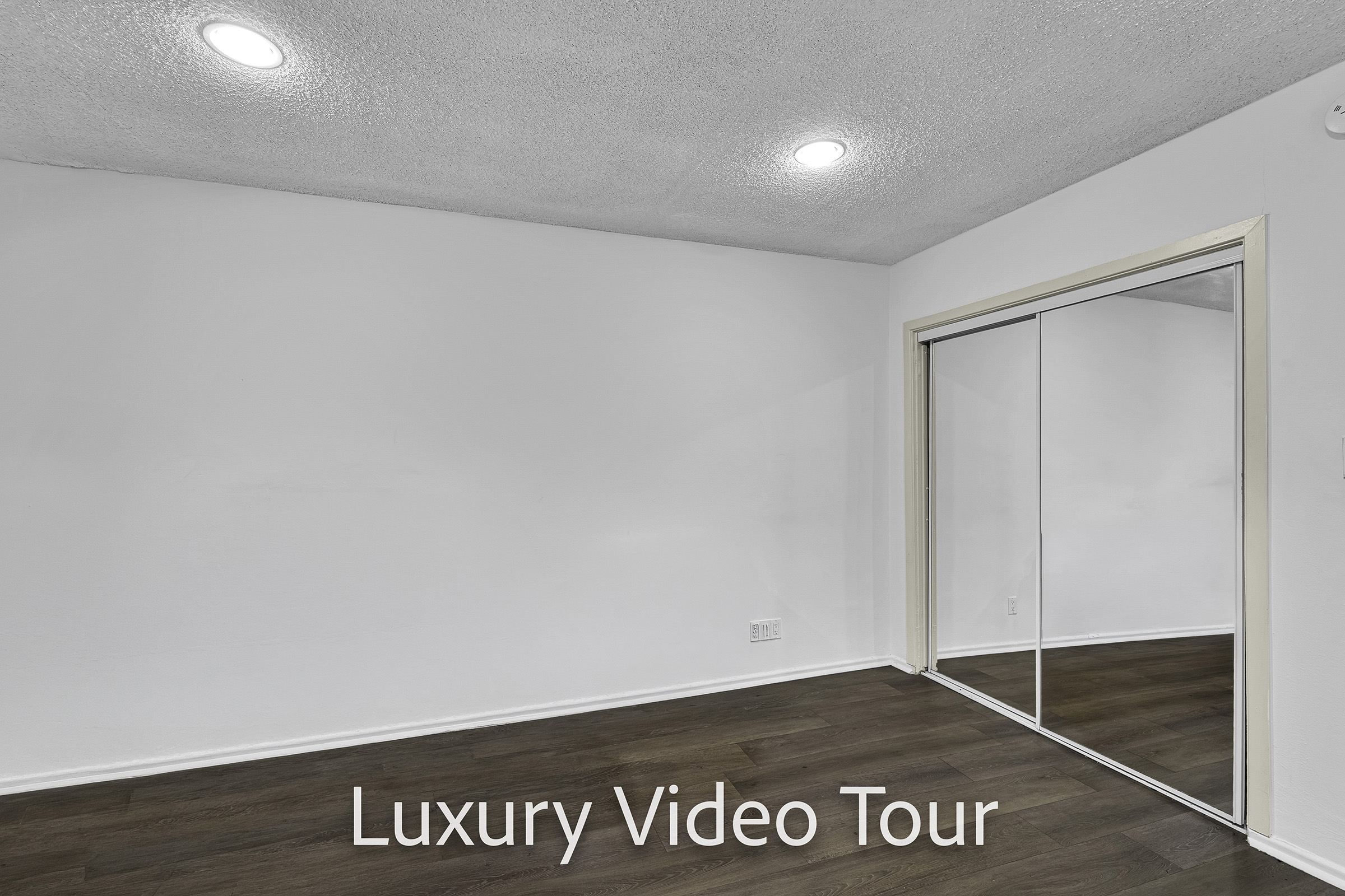 Empty interior space with light-colored walls and modern recessed lighting. A large mirror wardrobe reflects the bright room. The flooring is a rich, dark wood, contributing to a luxurious atmosphere. Text overlay reads "Luxury Video Tour."