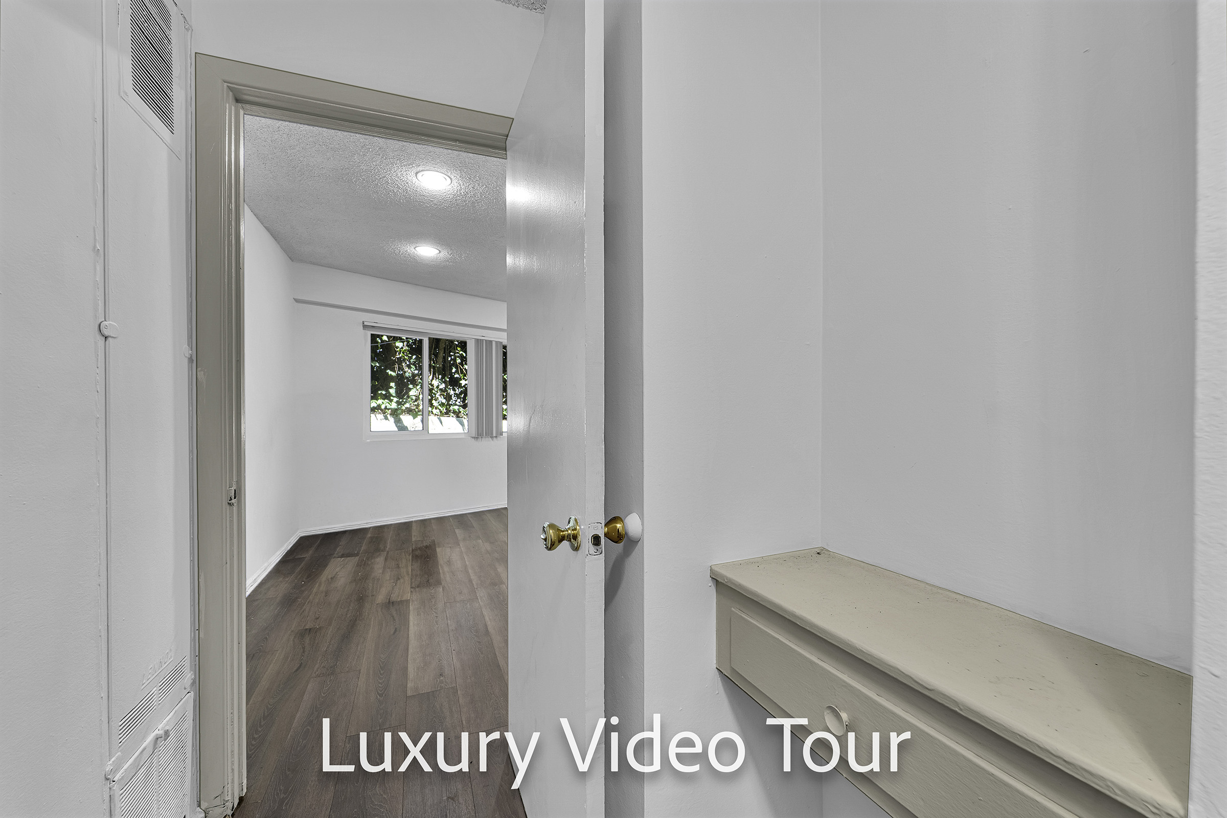 A doorway leads into a bright room with large windows, showcasing a modern interior. The walls are painted white, and the floor is a warm wood tone. A small ledge or shelf is seen next to the door, and the text "Luxury Video Tour" is prominently displayed in the foreground.