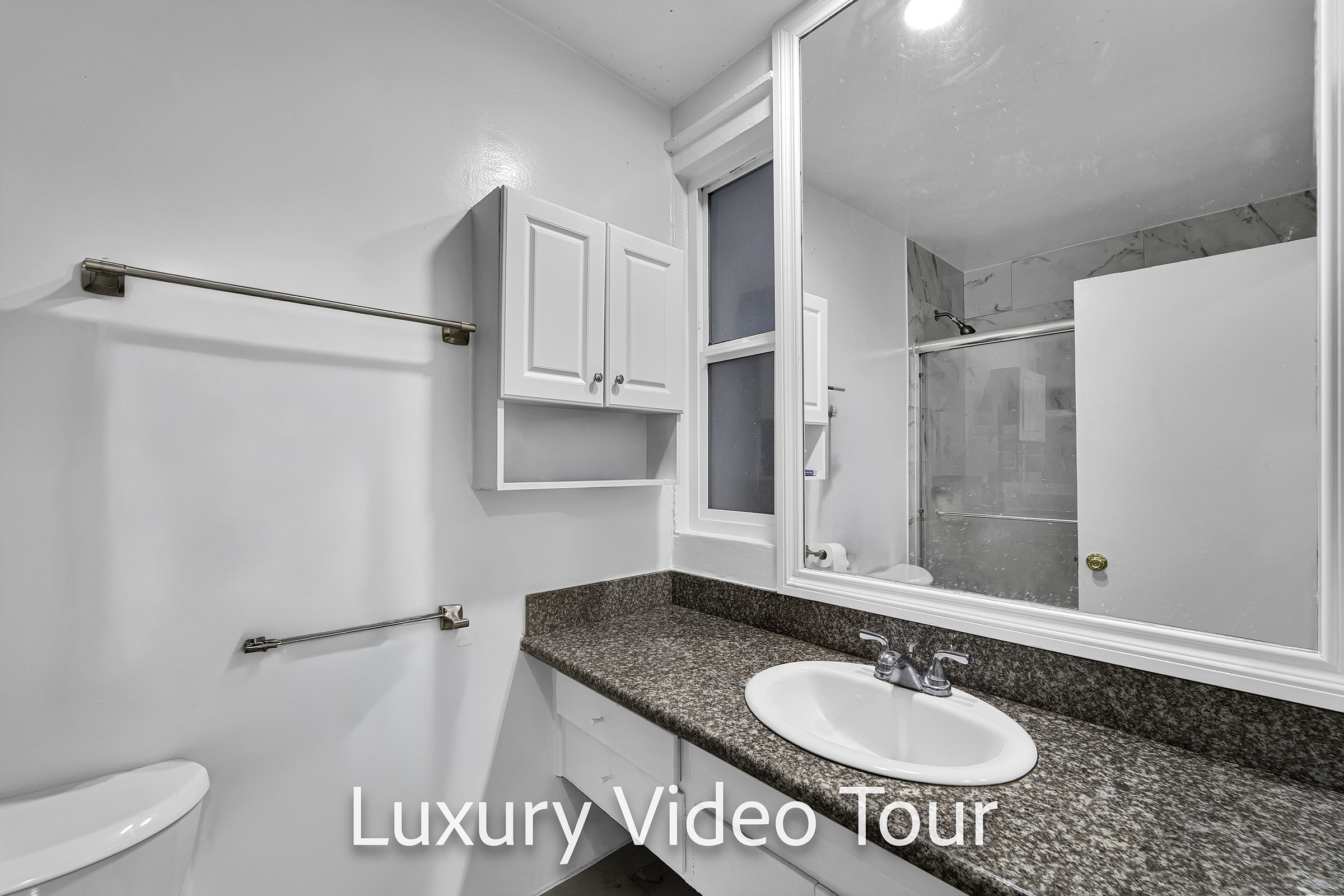 A modern bathroom featuring a white wall, a large mirror, a sink with a faucet, and a countertop. There is a shower area in the background with a frosted glass door, and a towel rack mounted on the wall. The space is well-lit and clean, showcasing a minimalist design.