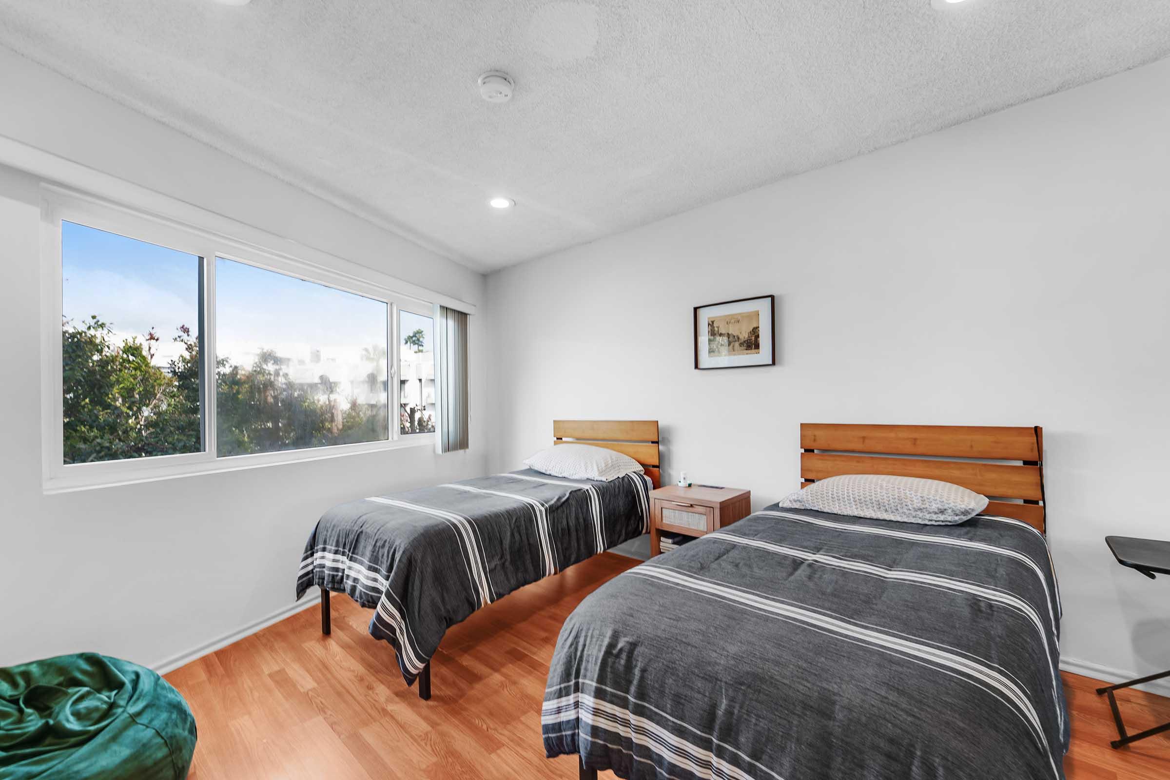 A bright, modern bedroom featuring two wooden twin beds with gray striped bedding. There's a small nightstand between the beds, and a window offers a view of greenery outside. The walls are white, and the flooring is light wood, creating a clean and inviting atmosphere.