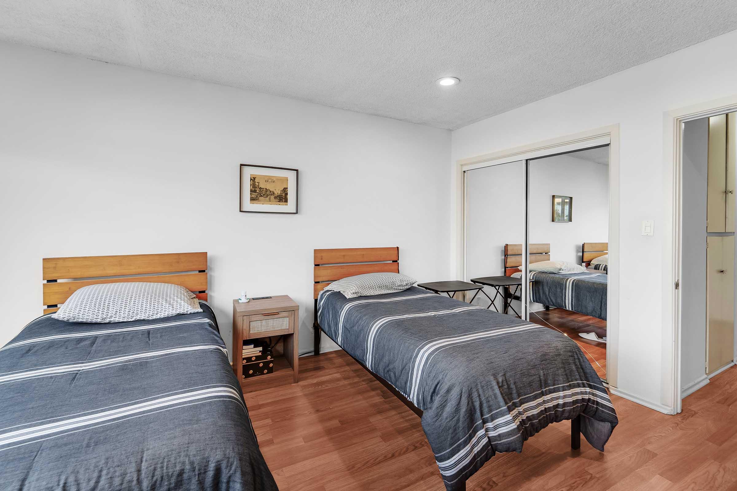 A bedroom featuring two single beds with striped bedding, a nightstand between them, and a framed picture on the wall. There is a large mirror reflecting the room and wooden flooring. The room has a bright and clean aesthetic with simple décor.