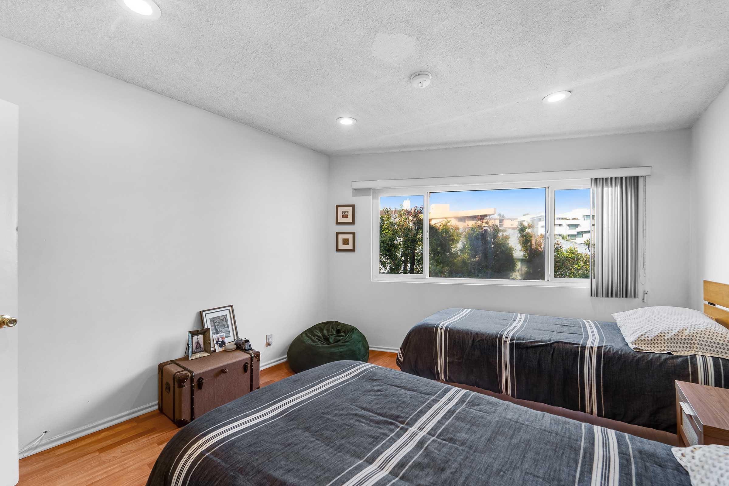 A cozy bedroom featuring two twin beds with striped bedding. There's a window with a view of greenery outside, a small nightstand with framed pictures, and a green bean bag chair. The walls are painted white, and the ceiling is textured. Natural light brightens the room.
