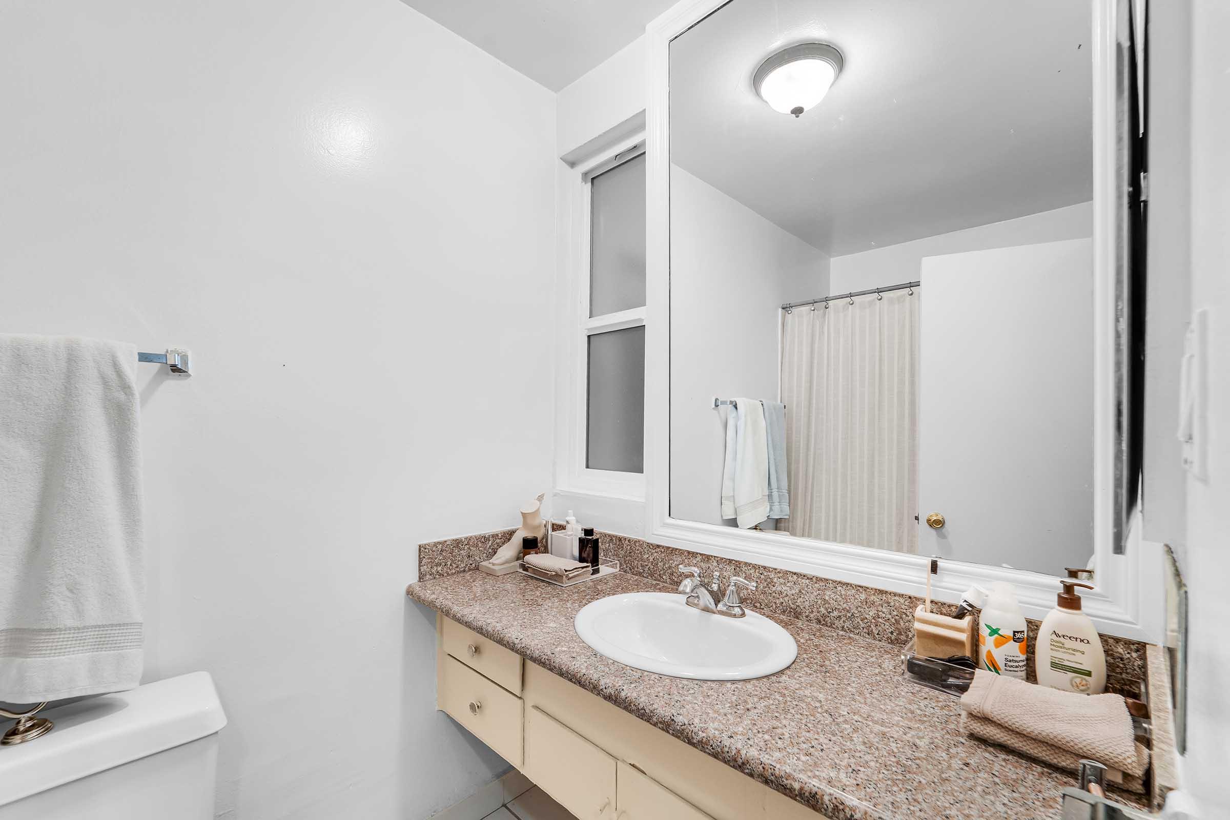 A clean, modern bathroom featuring a beige countertop with a sink, a mirror above it, and a shower curtain in the background. White towels hang on the wall, and various toiletries are neatly arranged on the counter. The walls are painted white, enhancing the bright and spacious feel of the room.