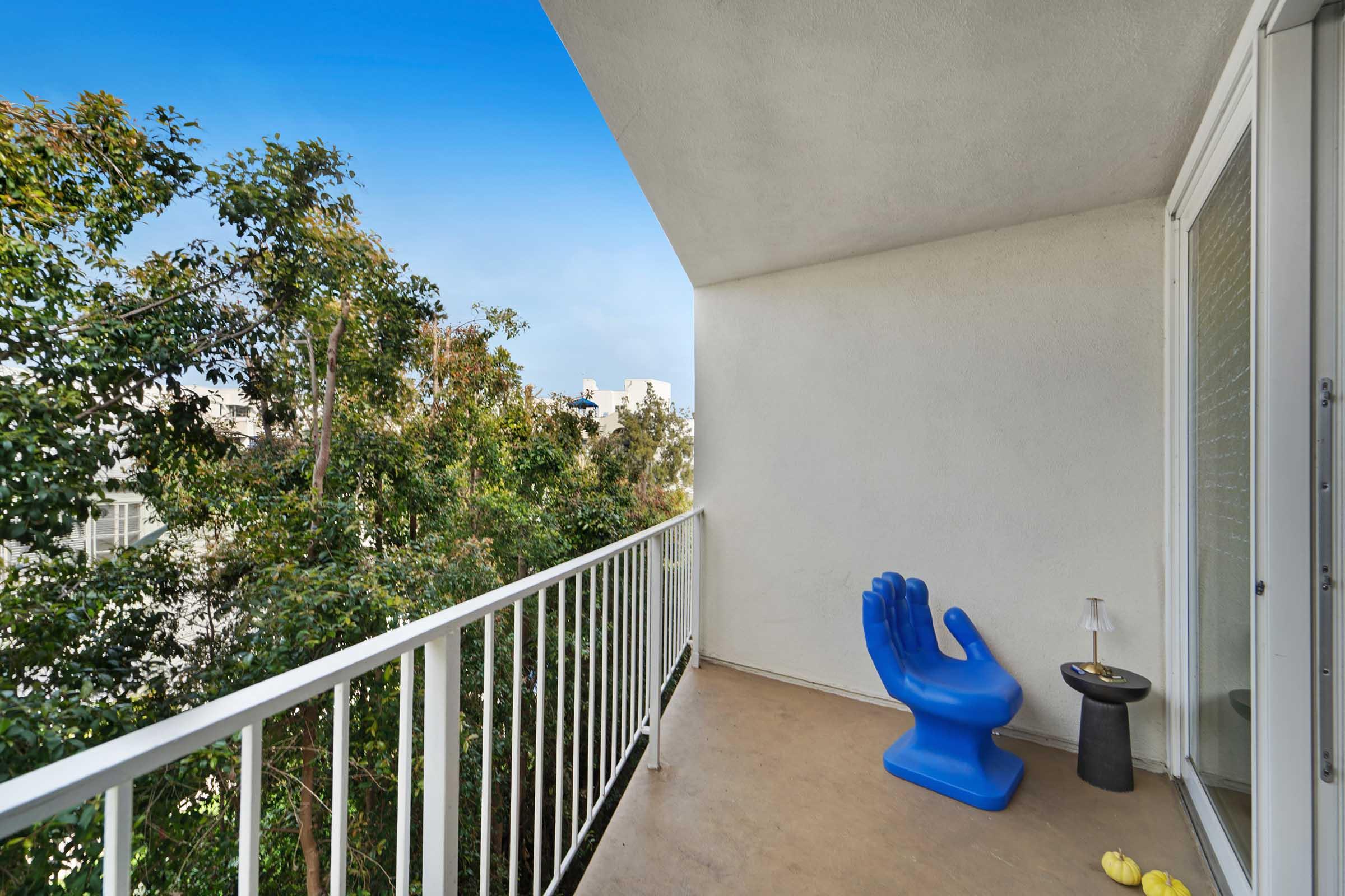 A balcony with a blue hand-shaped chair, a small black side table, and a lemon. The space is surrounded by greenery and features a clear blue sky in the background.