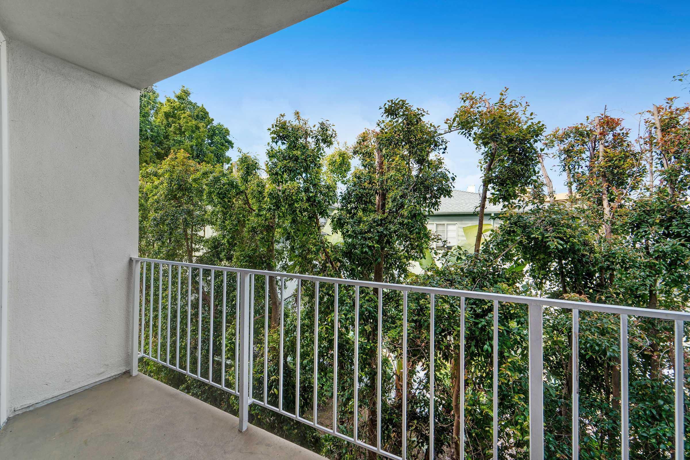 A balcony view with a white railing overlooking lush green trees and a bright blue sky. The space is well-lit and inviting, offering a peaceful outdoor area in an urban setting.